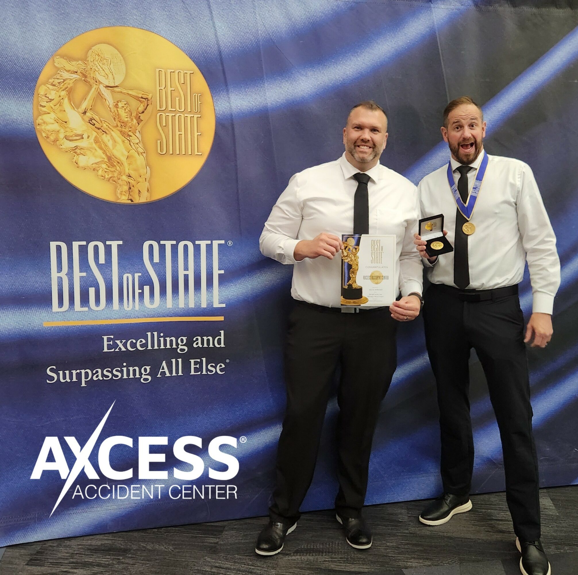 Two men in formal attire stand in front of a blue backdrop with awards, smiling and holding certificates and medals.