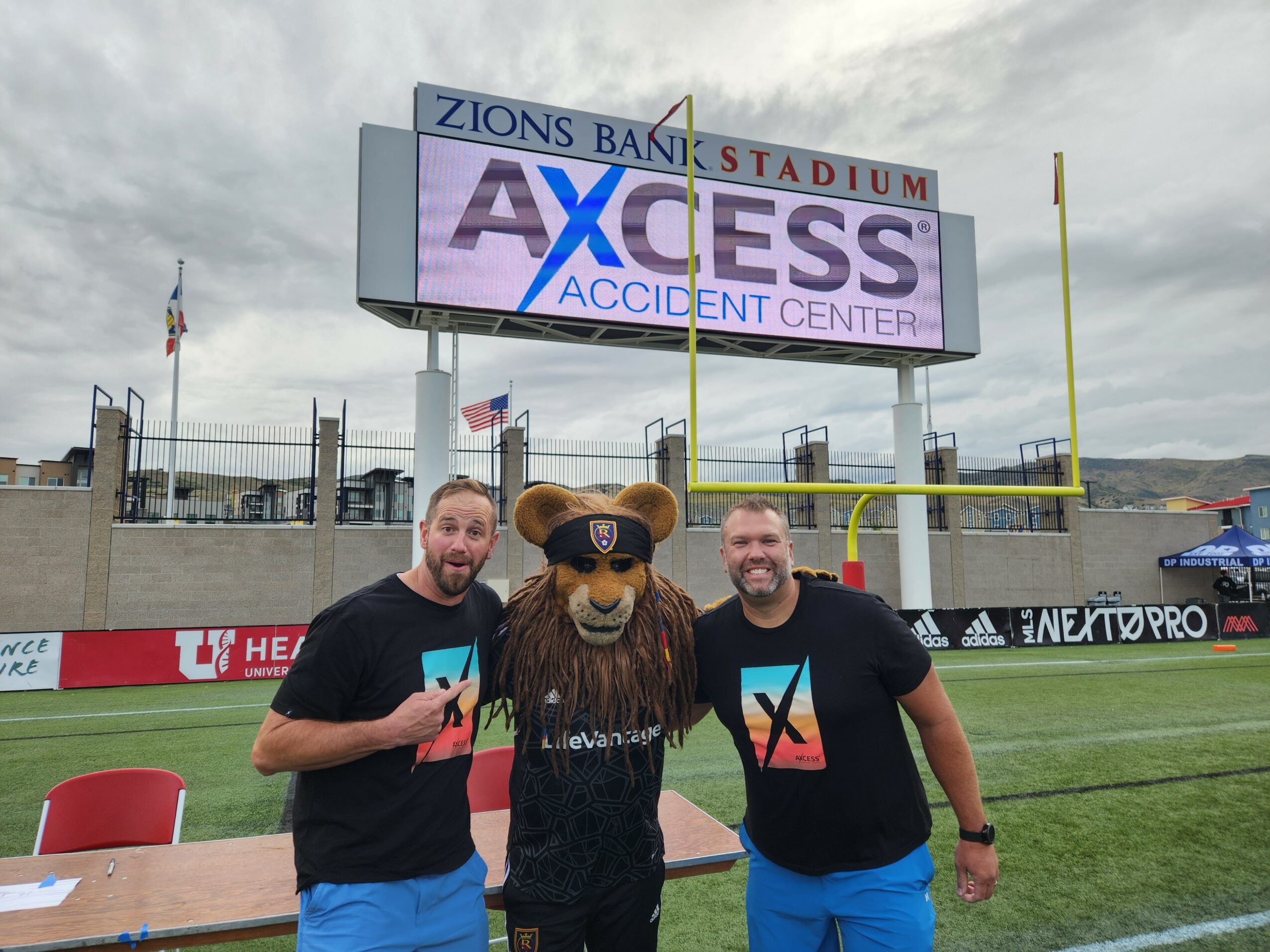 Two men stand on a football field with a bear mascot, smiling, in front of a scoreboard and stadium seating.