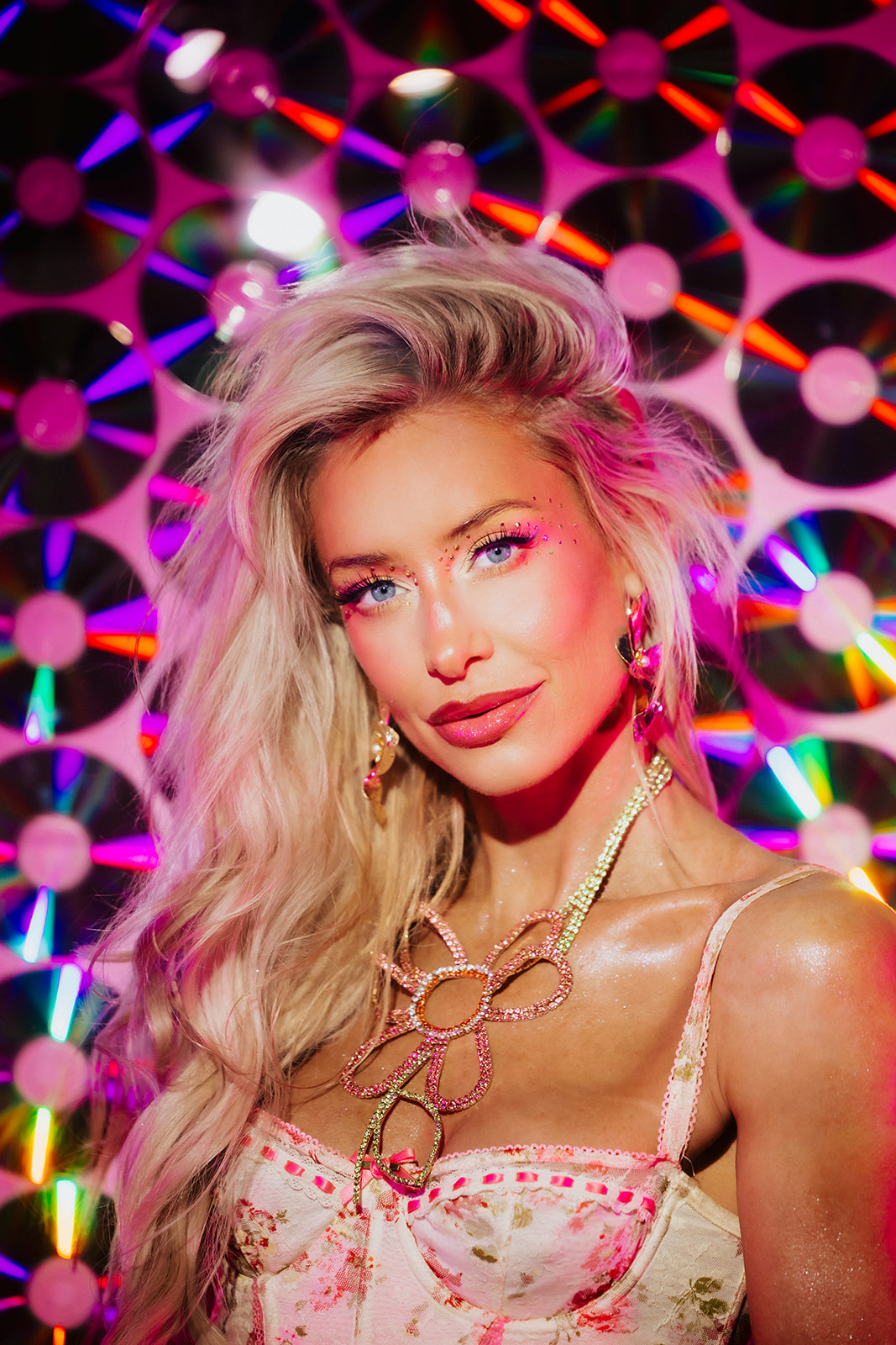 Young woman with long, wavy blonde hair and pink highlights, wearing jewelry, in front of a colorful, geometric background.