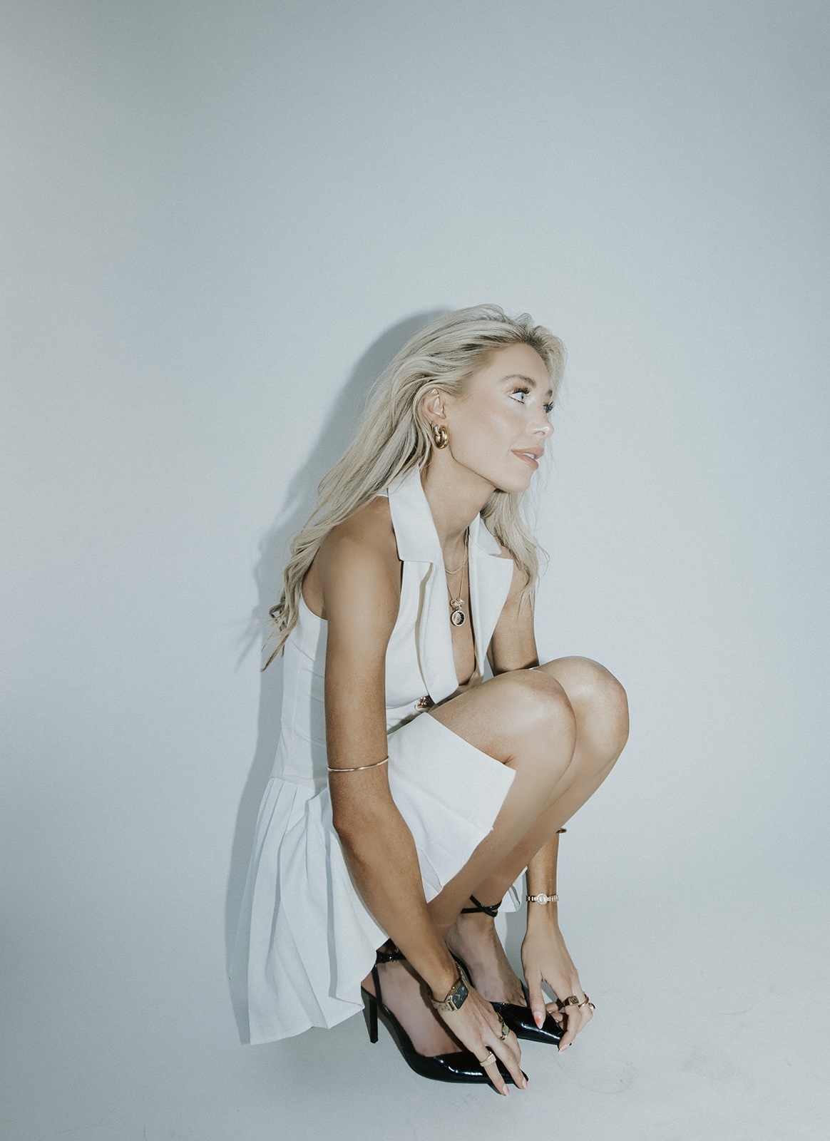 Woman with long blonde hair crouching against a plain light background, wearing a white dress and black high heels.