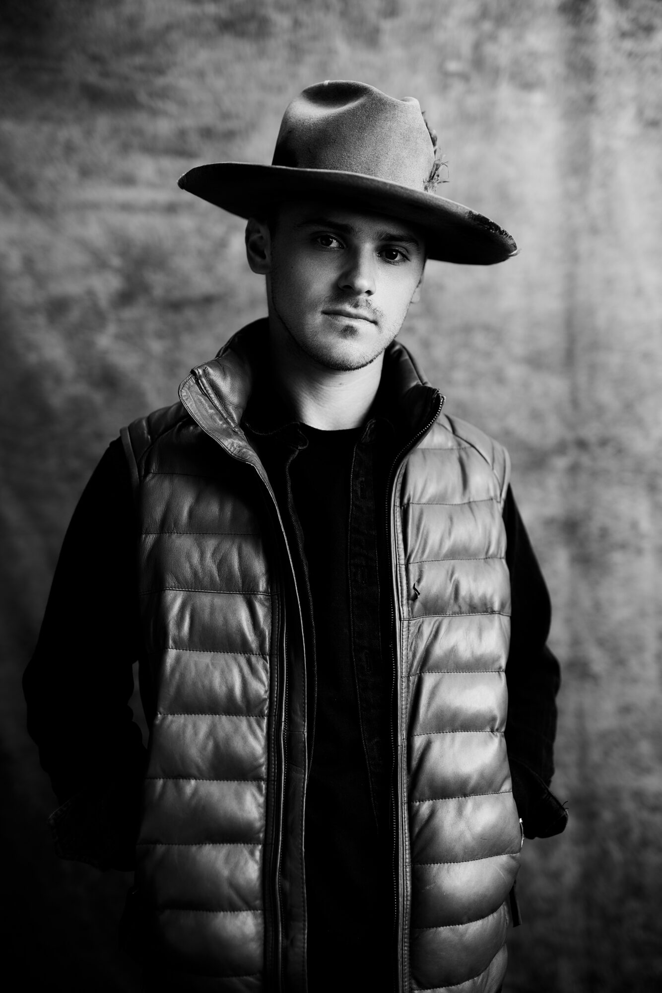 Young man wearing a wide-brimmed hat and puffer vest, standing against a textured background.