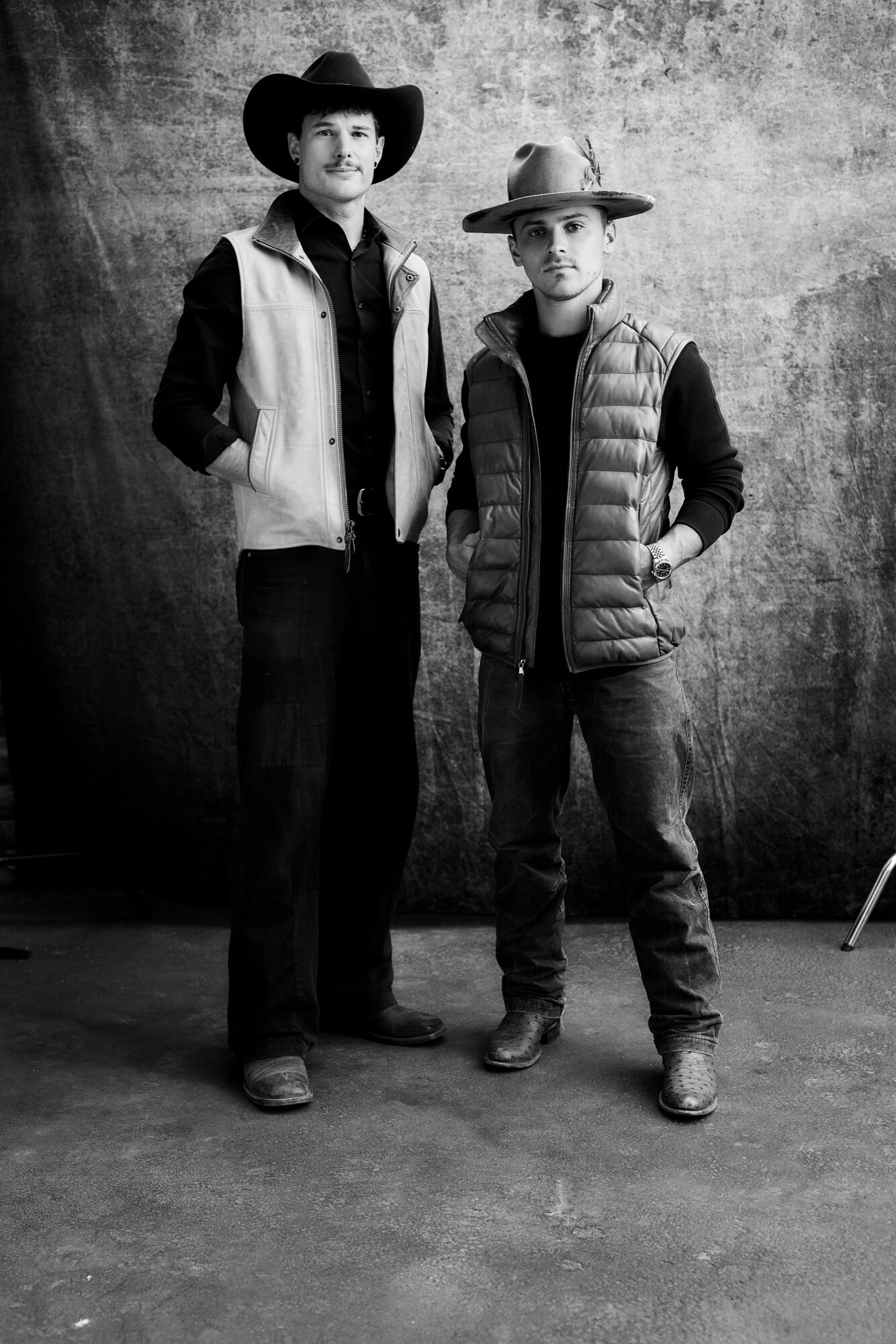 Two people standing side by side, one taller wearing a cowboy hat and vest, the other shorter with a hat and puffer jacket, against a textured background.