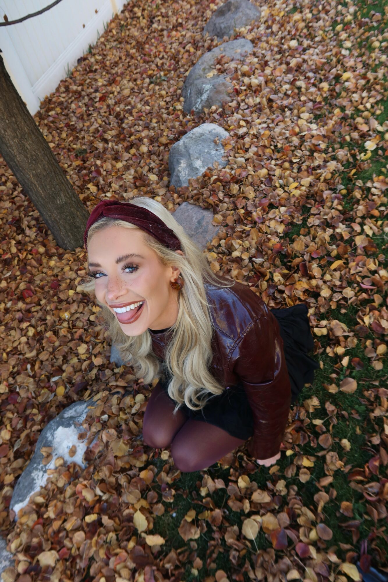 Woman with blonde hair smiling, kneeling among fallen autumn leaves, wearing a headband and maroon jacket.