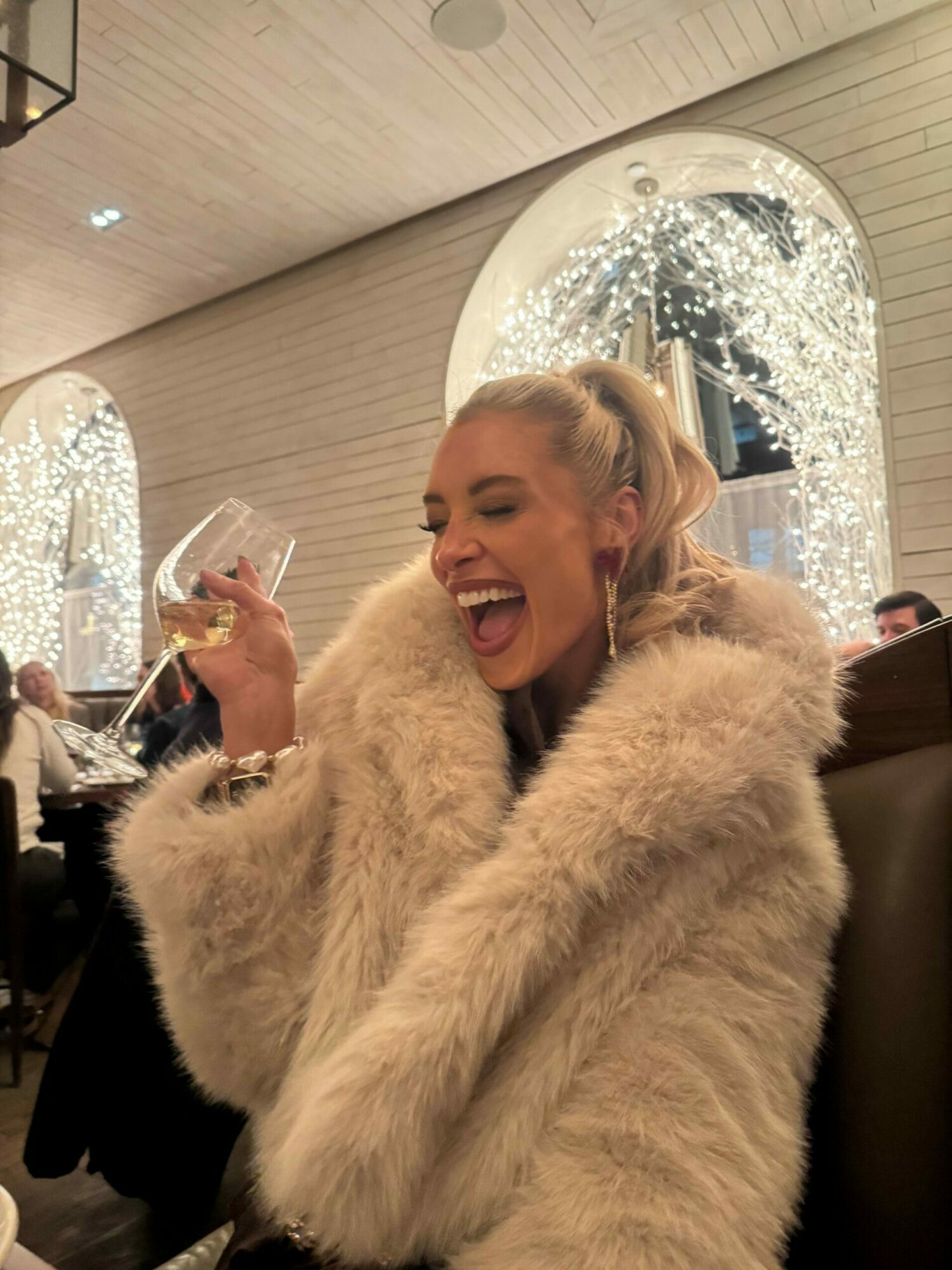 Person with blonde hair in a light-colored fur coat laughing and holding a glass, sitting indoors near a window with rain.