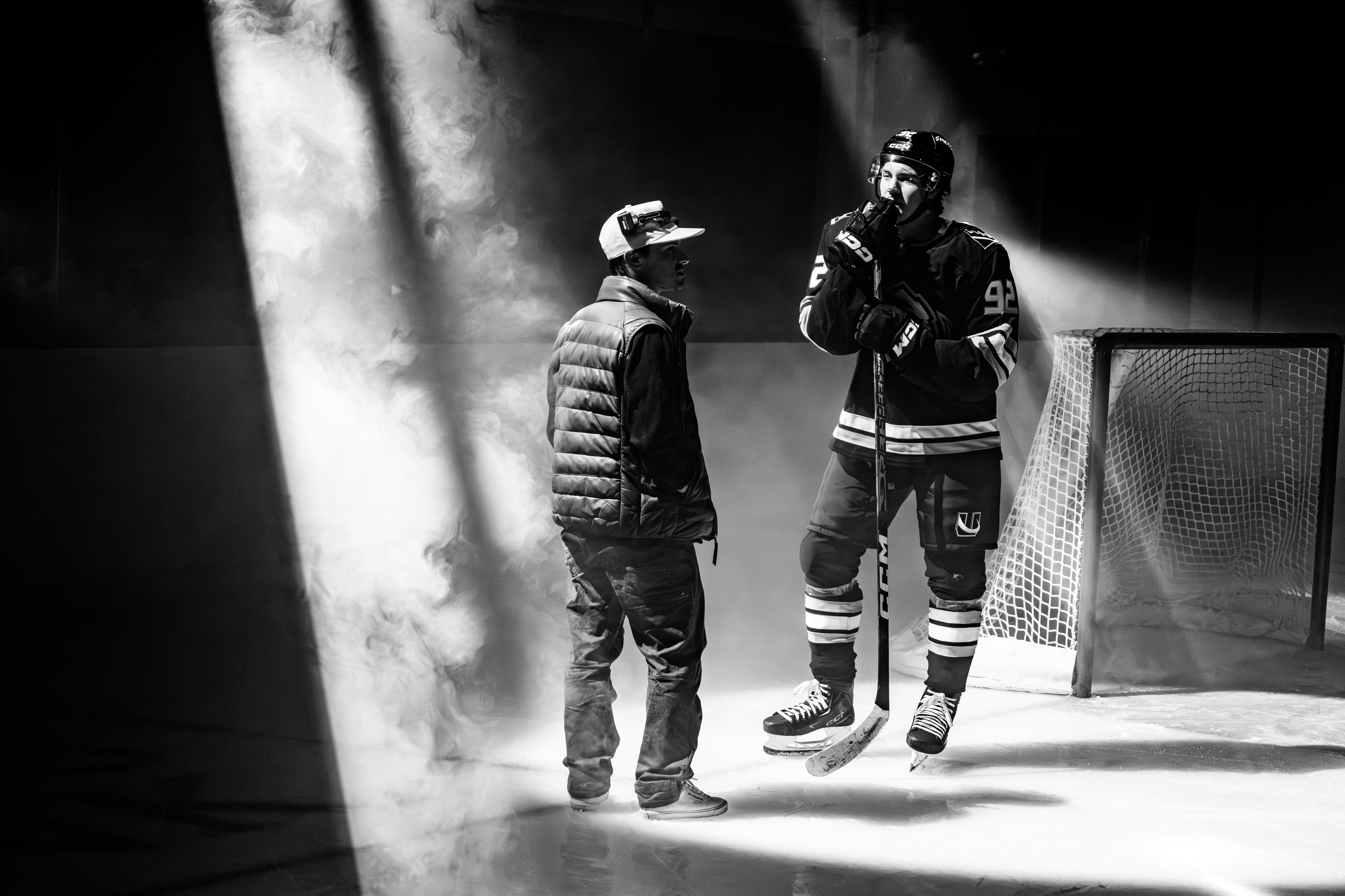 Two people, one in hockey gear, stand on ice rink with bright light and fog effects, talking near a goal net.