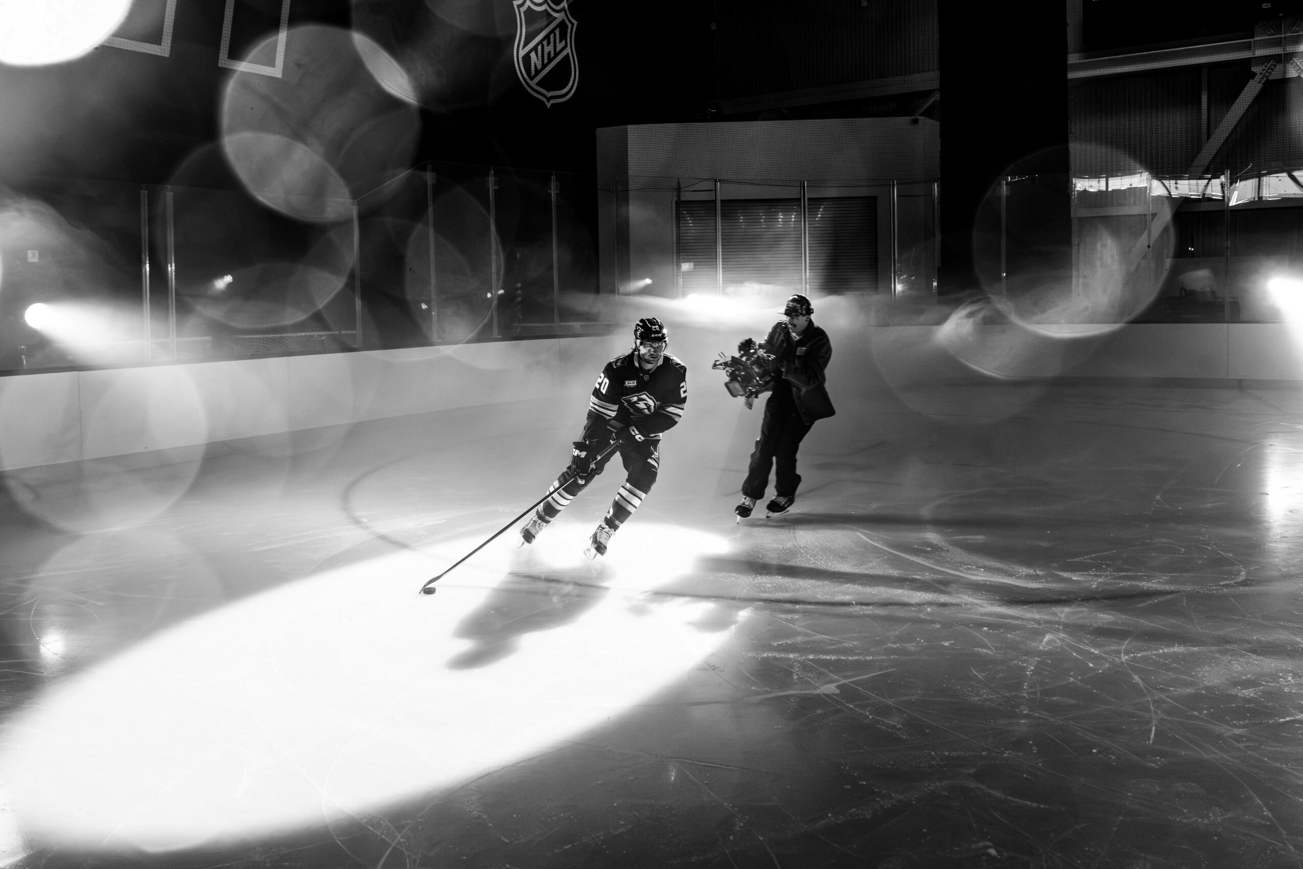 Two people ice skating indoors, one skating forward and the other behind, with bright lights and reflections on the ice.