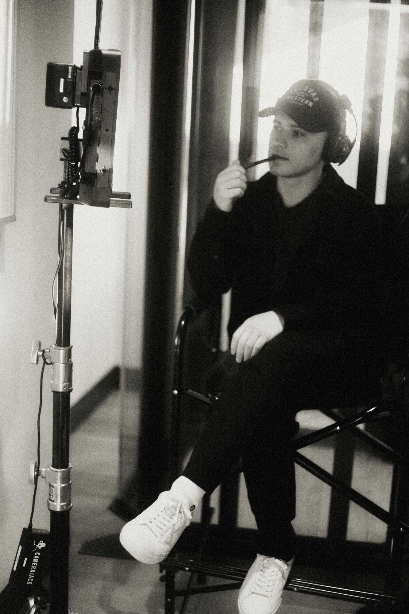 Person sitting on a chair, wearing a cap and white sneakers, with a mirror and vertical blinds in the background.