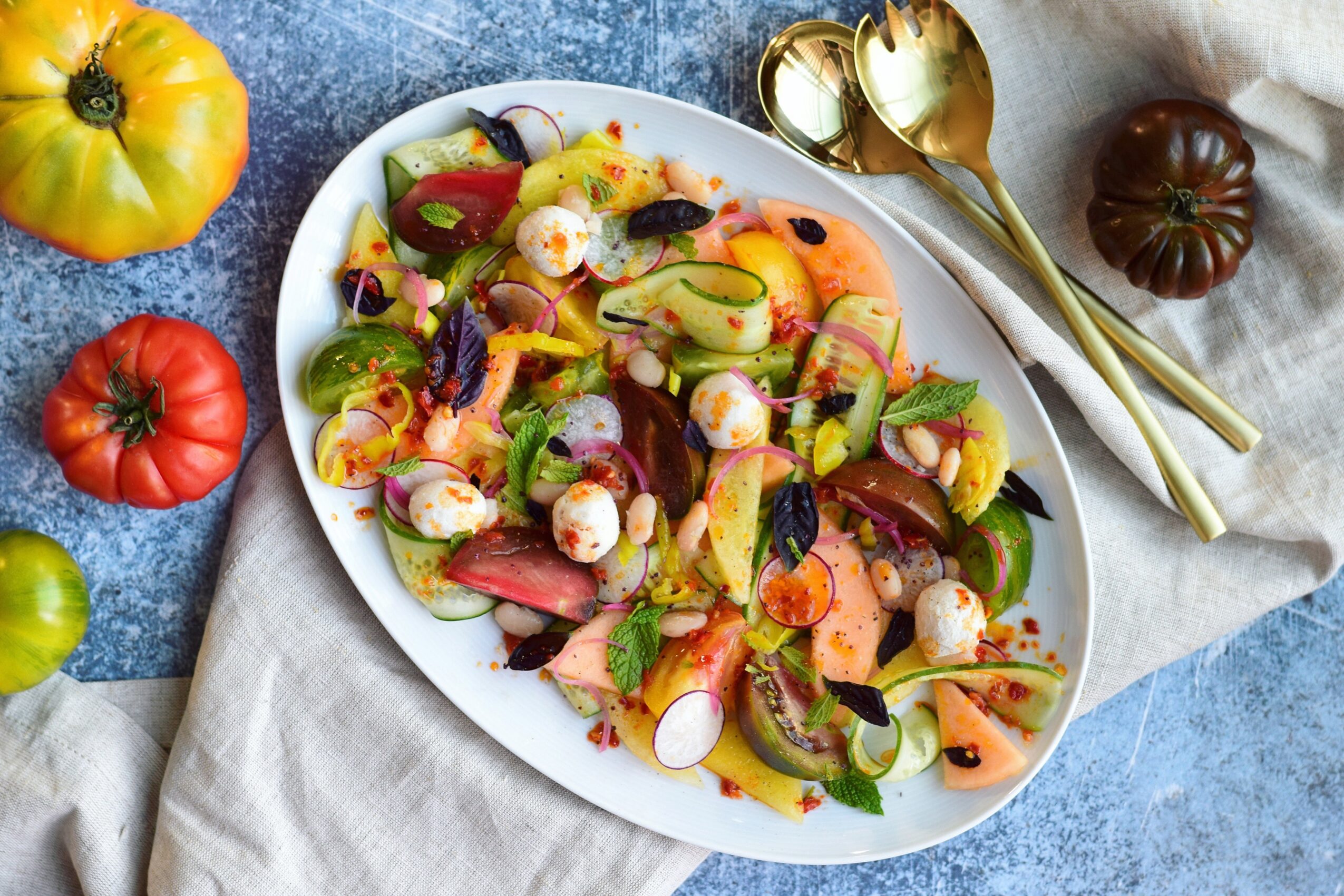 Oval platter of colorful salad with sliced vegetables, herbs, and round white pieces, surrounded by tomatoes and serving spoons.