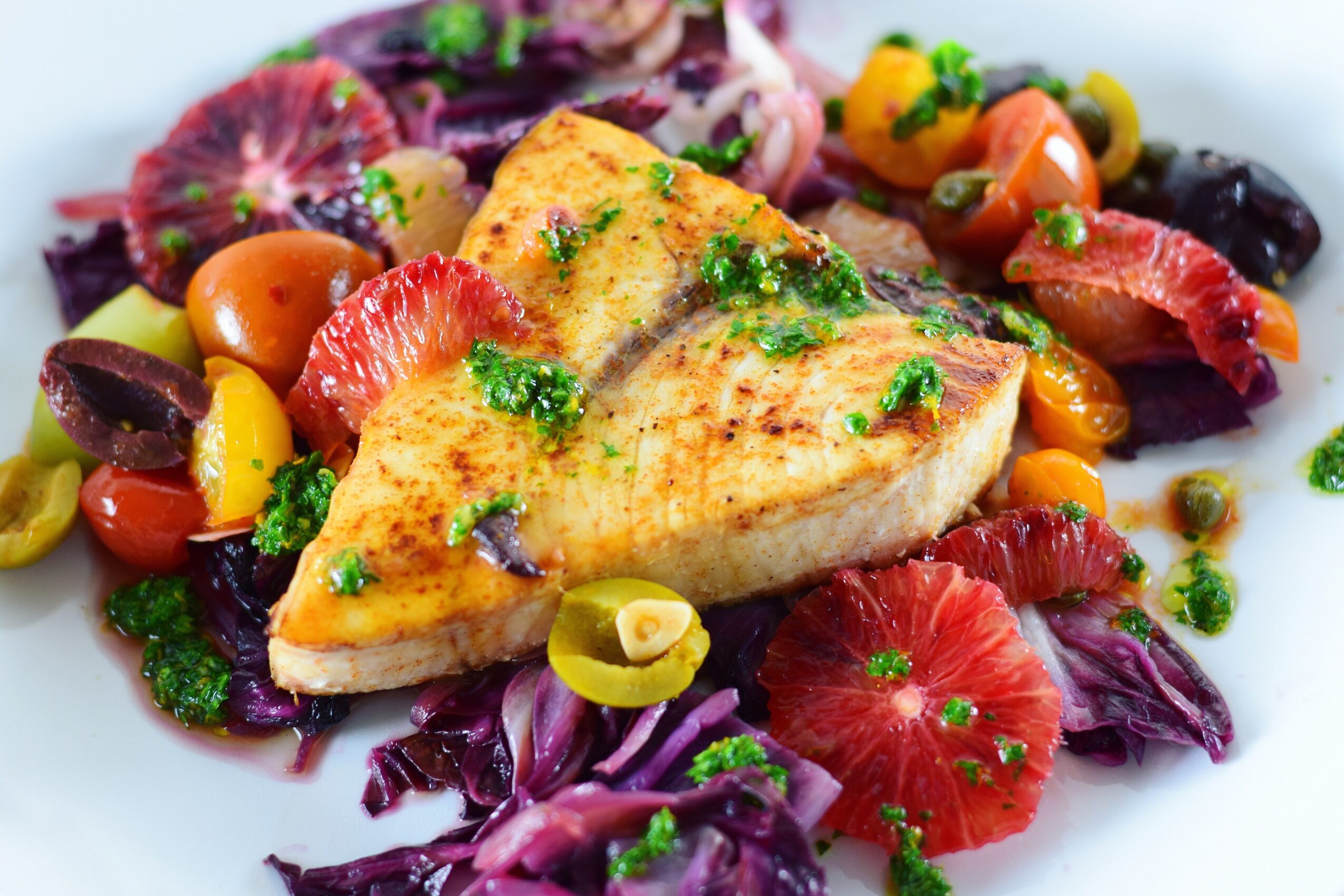 Grilled fish fillet surrounded by citrus segments, olives, tomatoes, peppers, and leafy greens on a white plate.