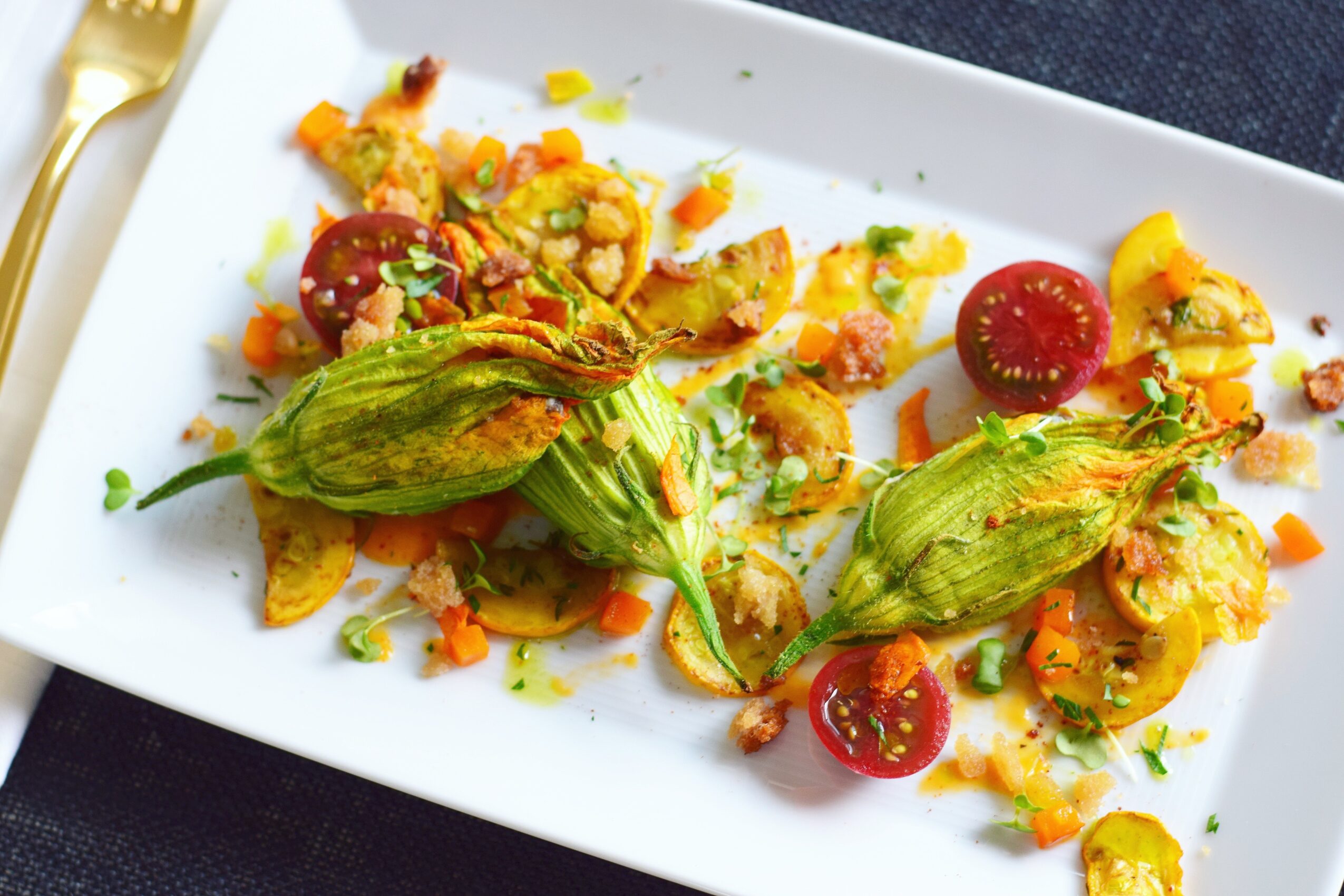 A white rectangular plate with stuffed squash blossoms, chopped vegetables, herbs, and halved cherry tomatoes.