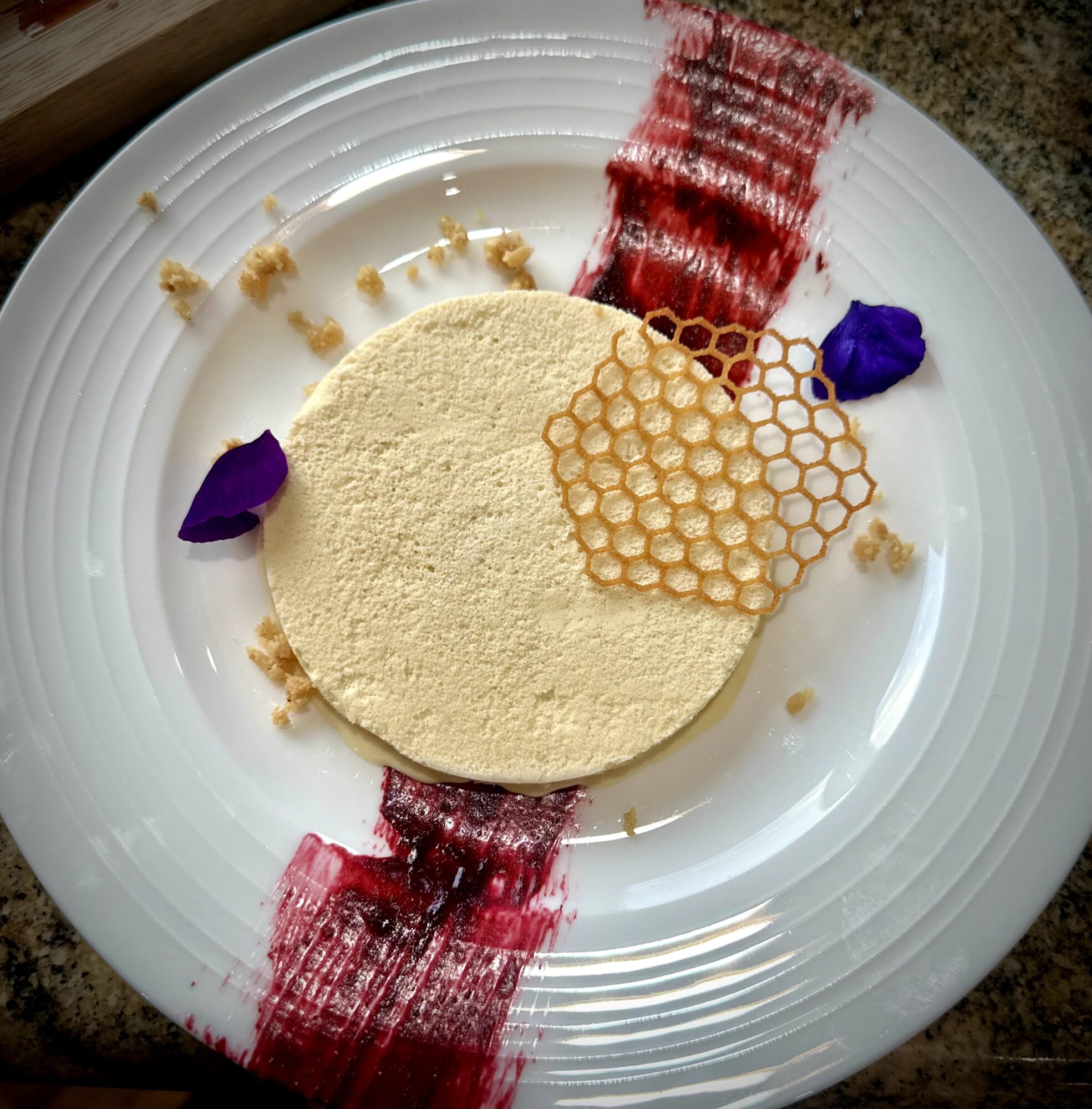 Round beige dessert on a white plate, topped with honeycomb, red sauce, and purple petals.