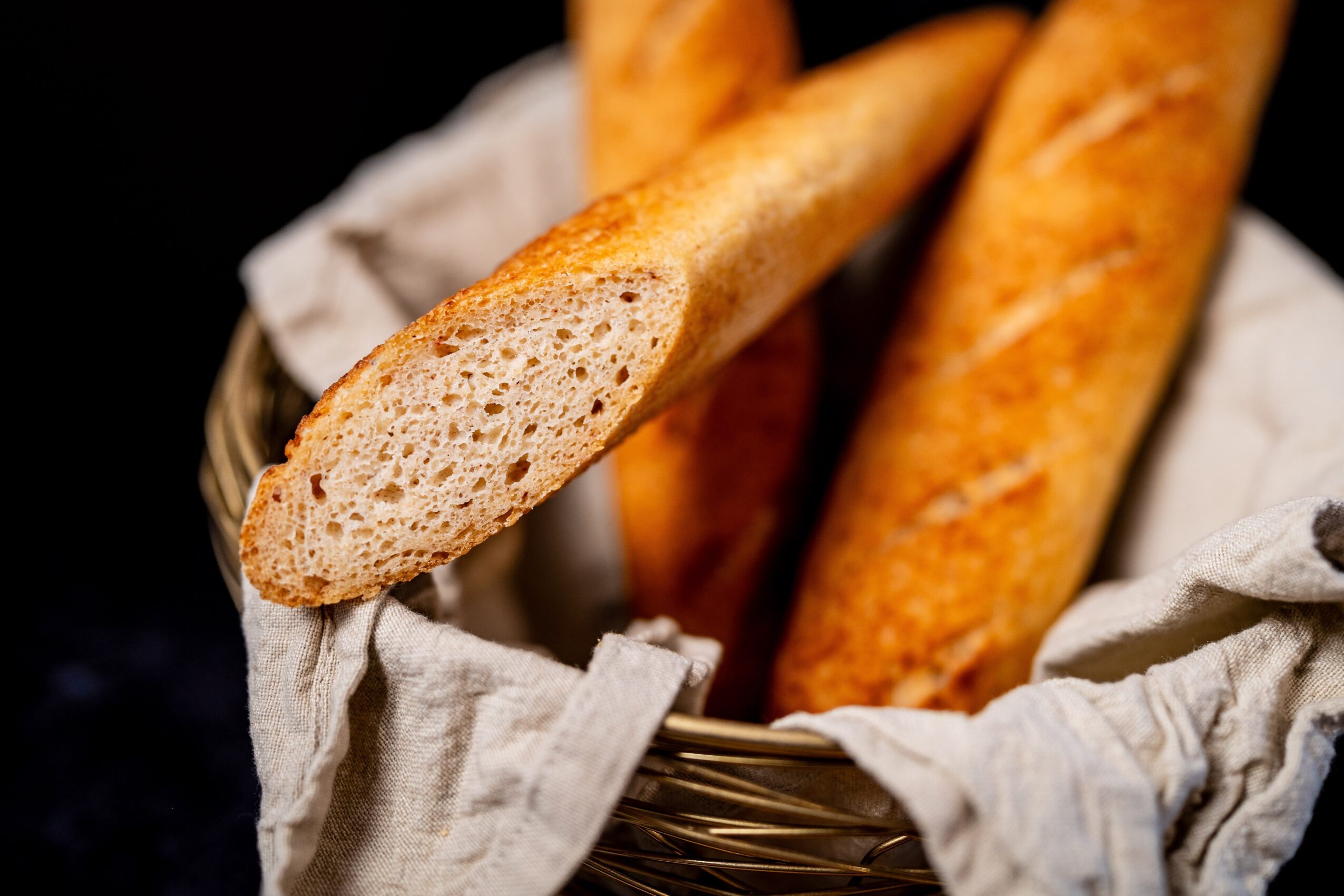 Several golden-brown baguettes rest in a cloth-lined basket; one sliced end reveals its airy interior.