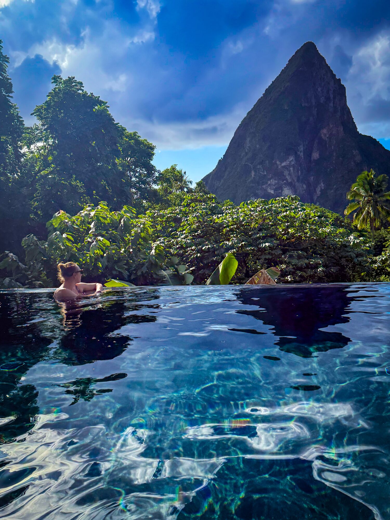 A person swims in a clear pool beneath a steep mountain, with lush trees and a cloudy blue sky.