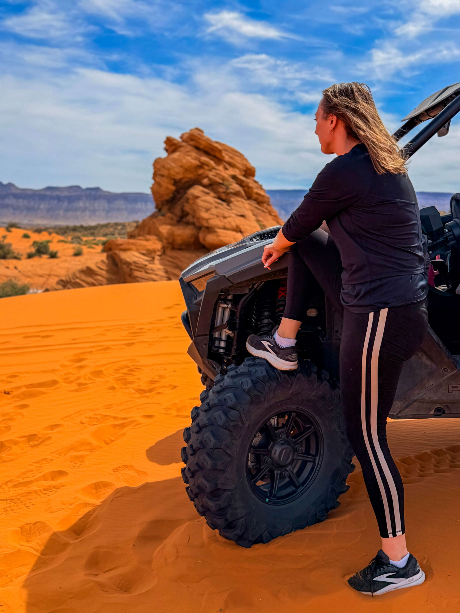 A person sits on an off-road vehicle in orange desert sand, with a rock formation and distant mountains behind them.