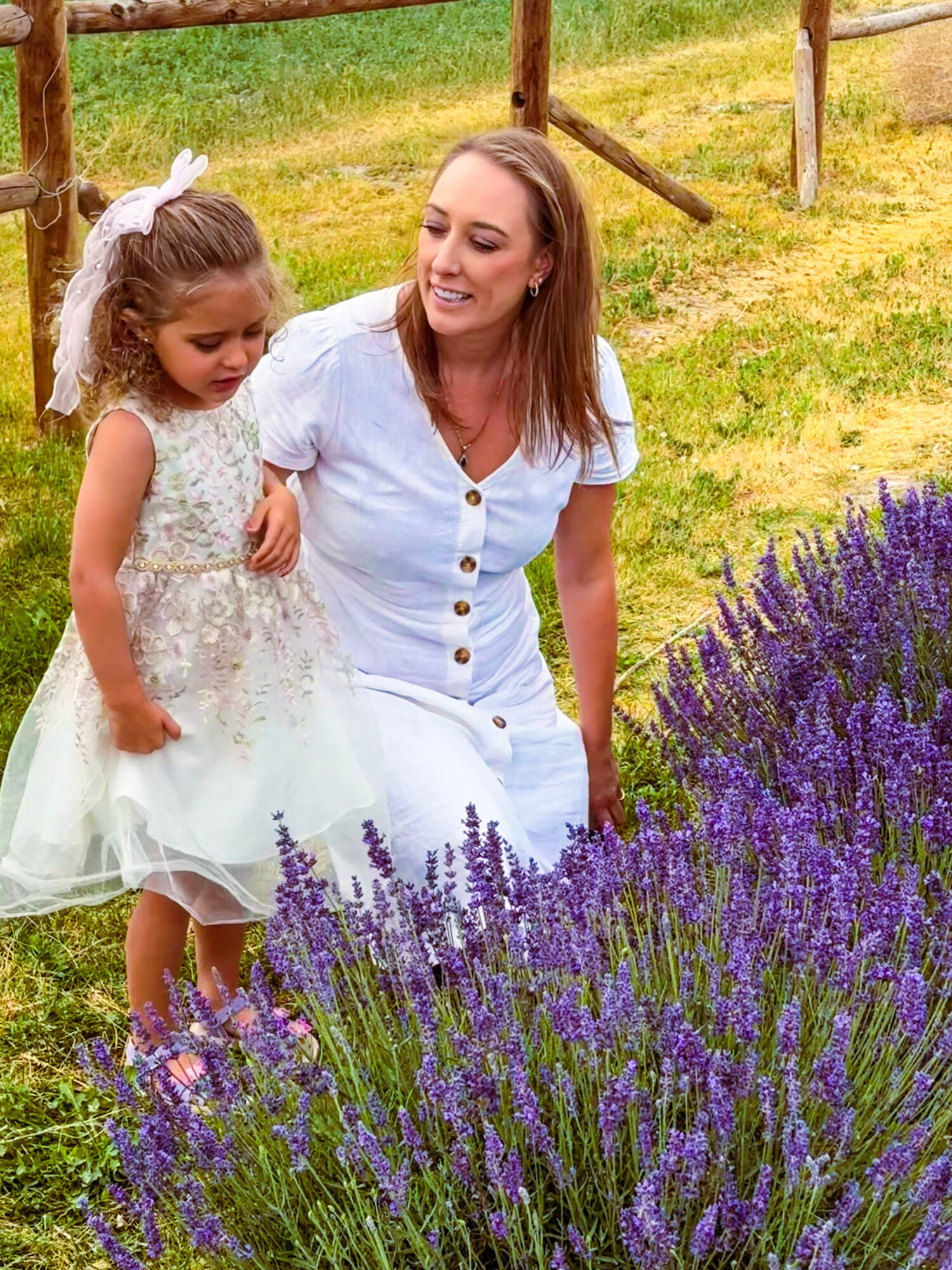 A woman and young child stand beside purple flowering plants in a grassy field.