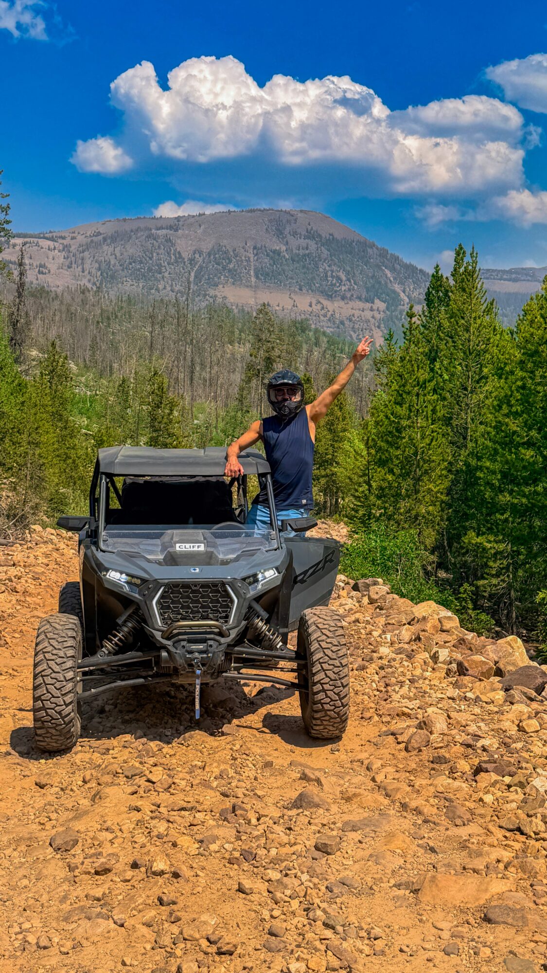 A person in an off-road vehicle raises an arm on a rocky trail, with trees and a mountain behind them.