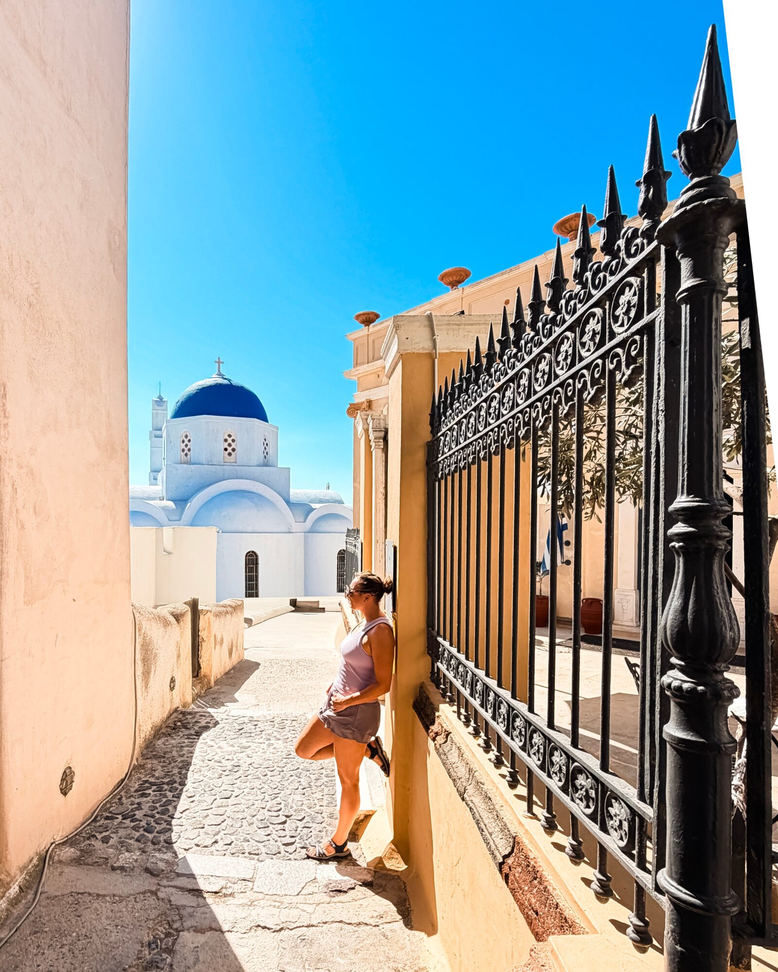 A person stands beside an ornate black iron gate on a sunlit walkway, with a blue-domed building in the background.