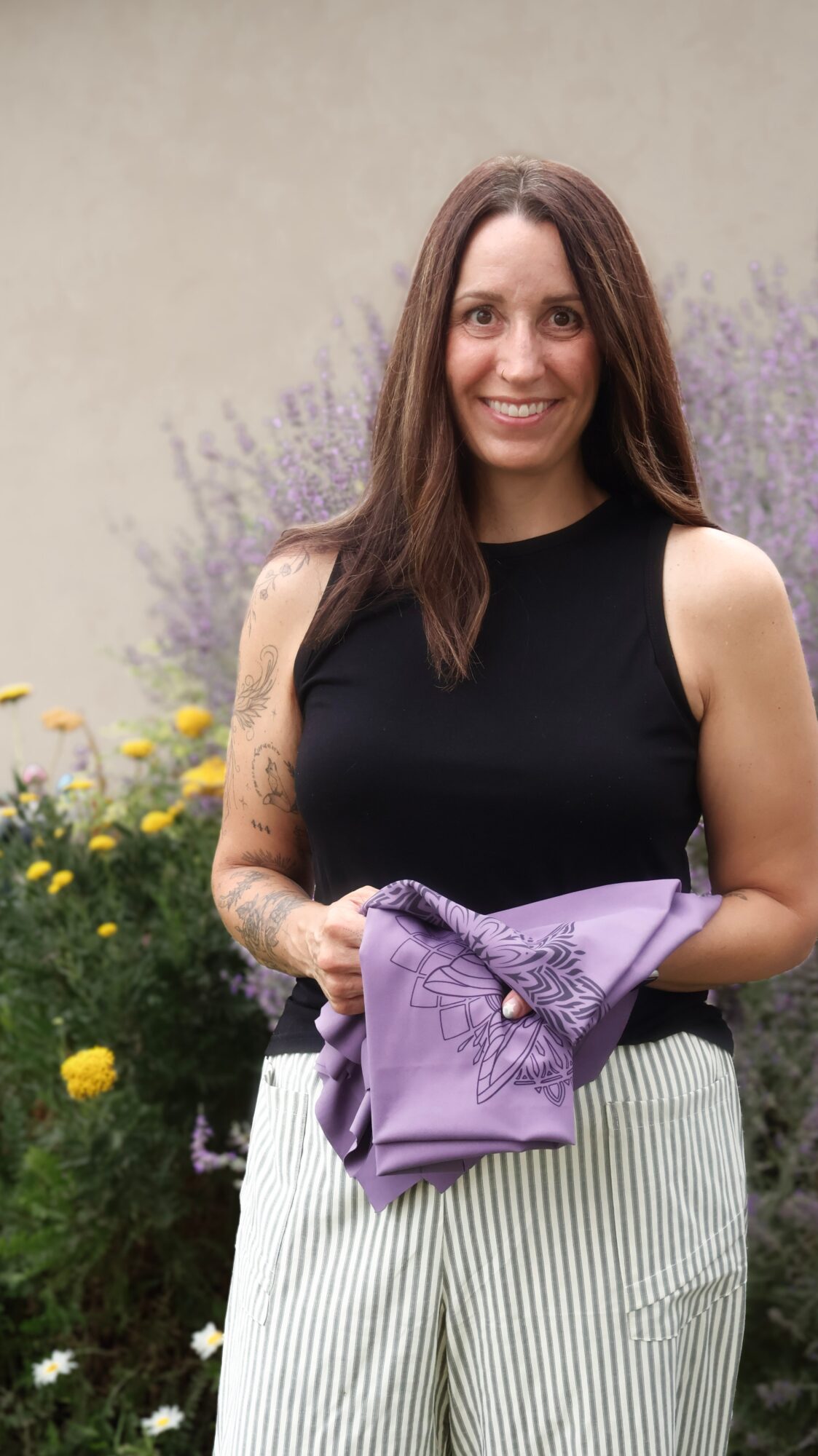 A person standing in a garden holds a folded purple garment, wearing a black top and striped pants.