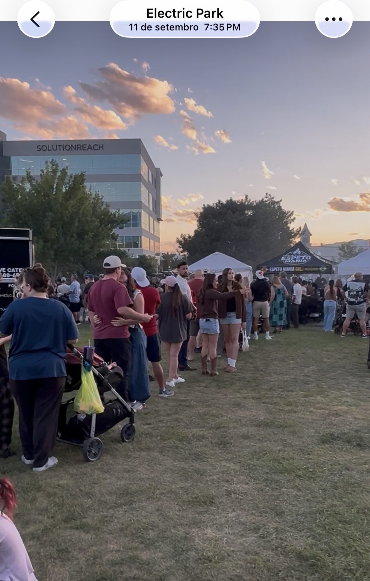 People queue on a grassy field near tents and a building beneath a sunset sky.