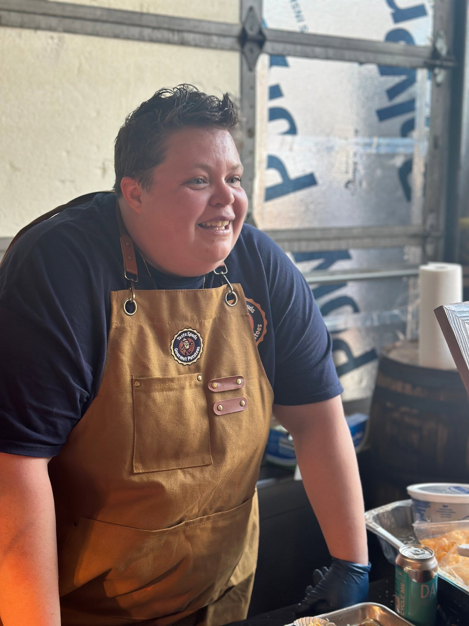 A person wearing a brown apron leans on a counter inside a workshop or food-service space.