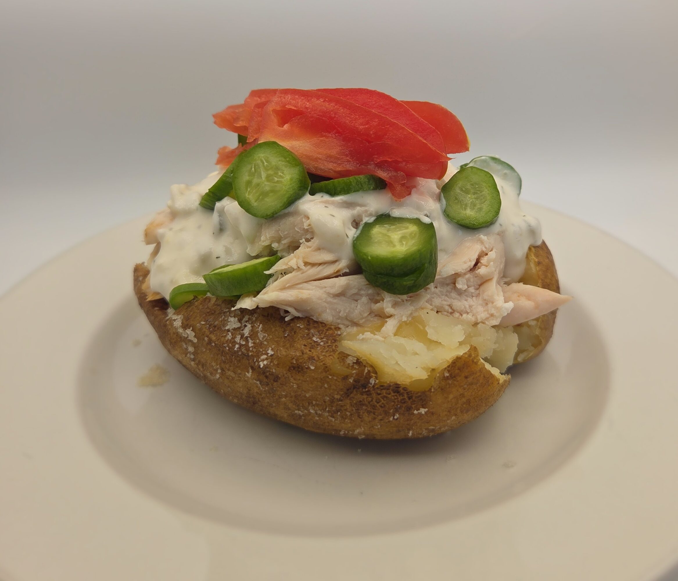 Baked potato topped with shredded chicken, creamy sauce, sliced green vegetables, and red tomato.