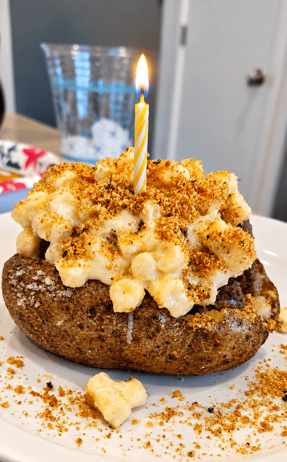 A baked potato topped with macaroni and cheese, seasoning, and a lit striped candle on a plate.
