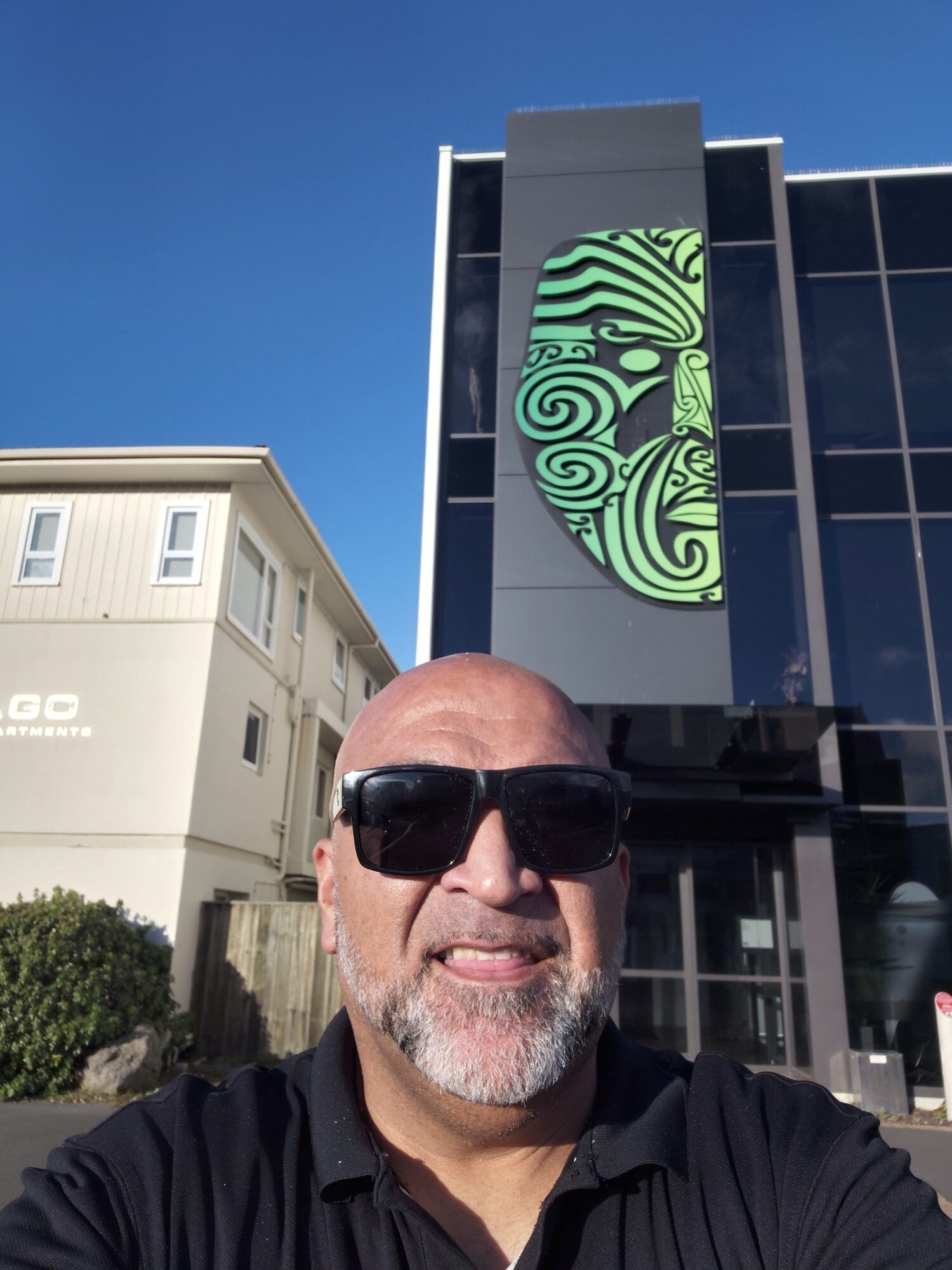 A bald person wearing sunglasses stands beside a modern building with a green patterned sign.