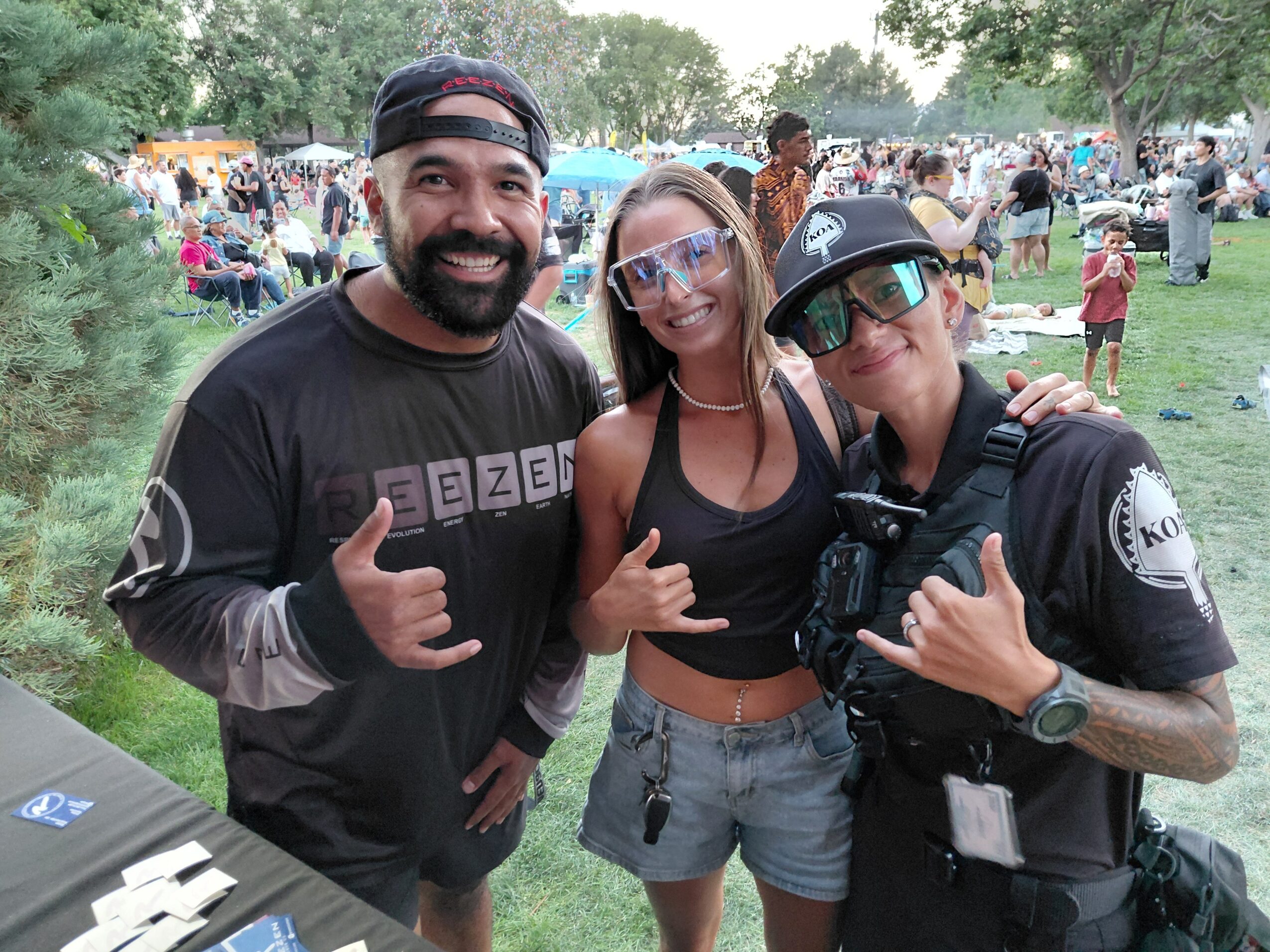 Three people pose together at an outdoor event, with a crowd and trees in the background.
