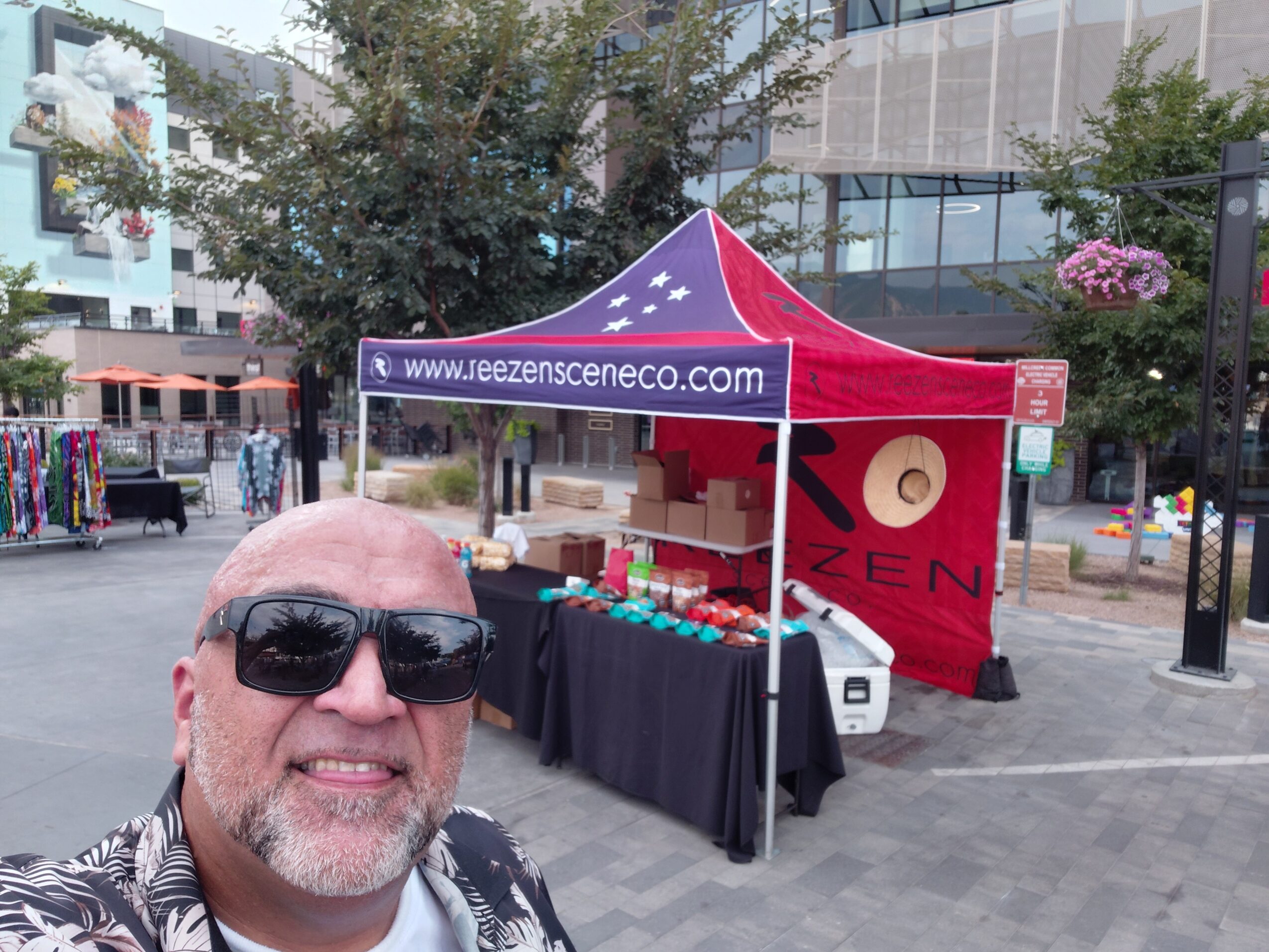 Person wearing sunglasses takes a selfie beside a red market tent with products on a city plaza.