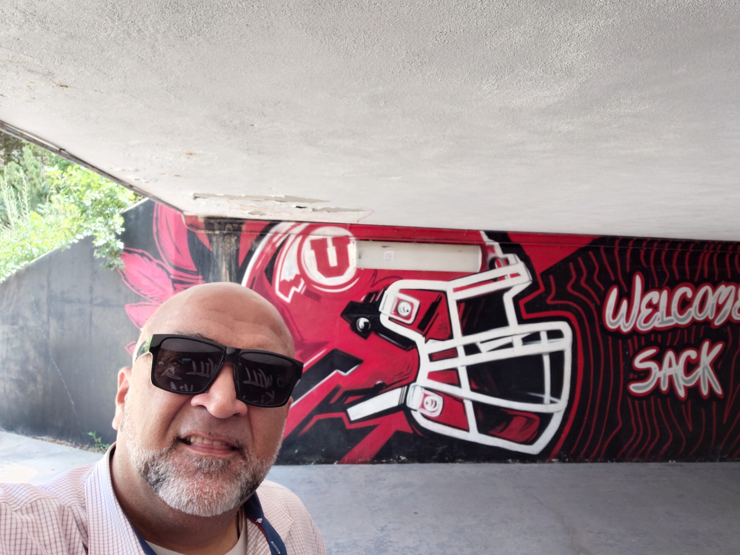 A man wearing sunglasses poses before a red-and-black mural of a football helmet and welcome text.