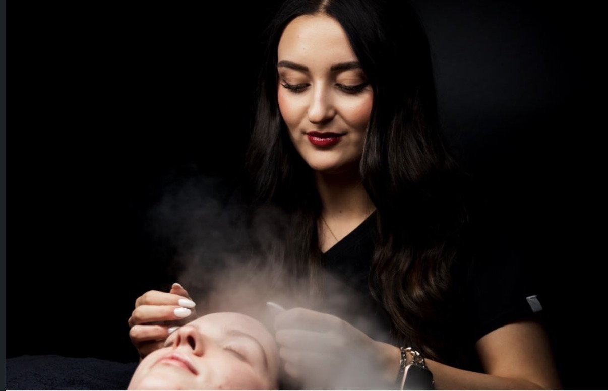 A person applies a steaming facial treatment to another person lying down.