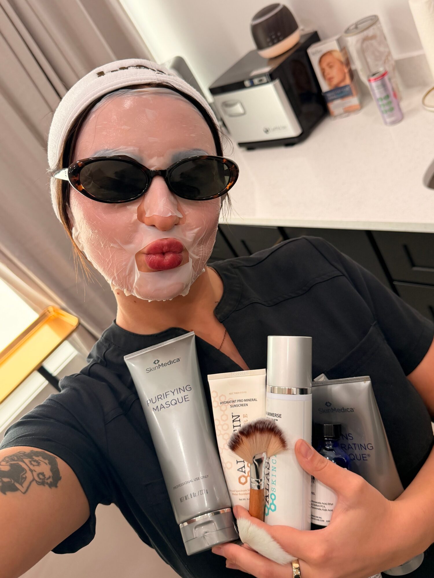 Person wearing a white facial mask and sunglasses holds several skincare product tubes beside a counter.
