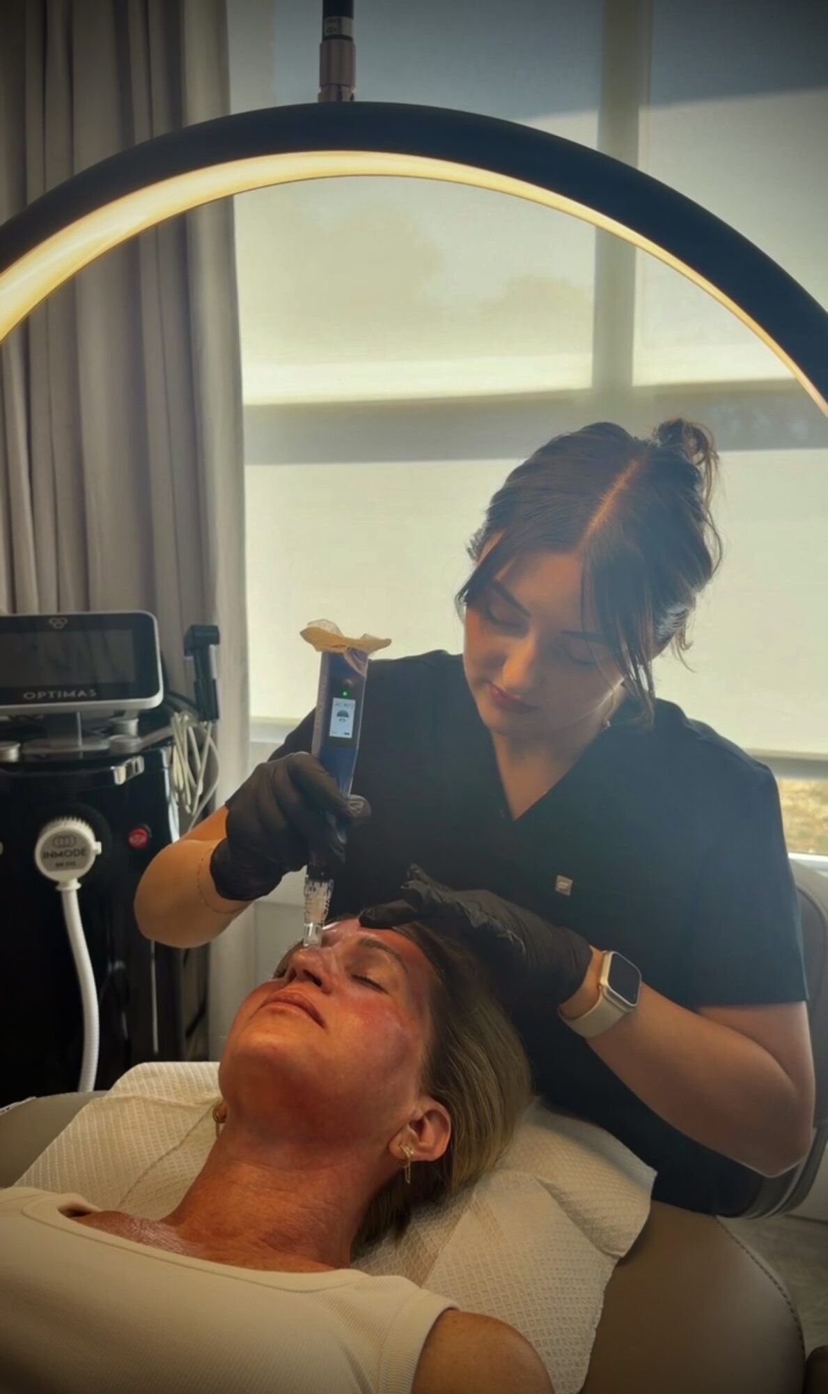 A person reclines with eyes closed while another person holds a handheld device near their face in a treatment room.