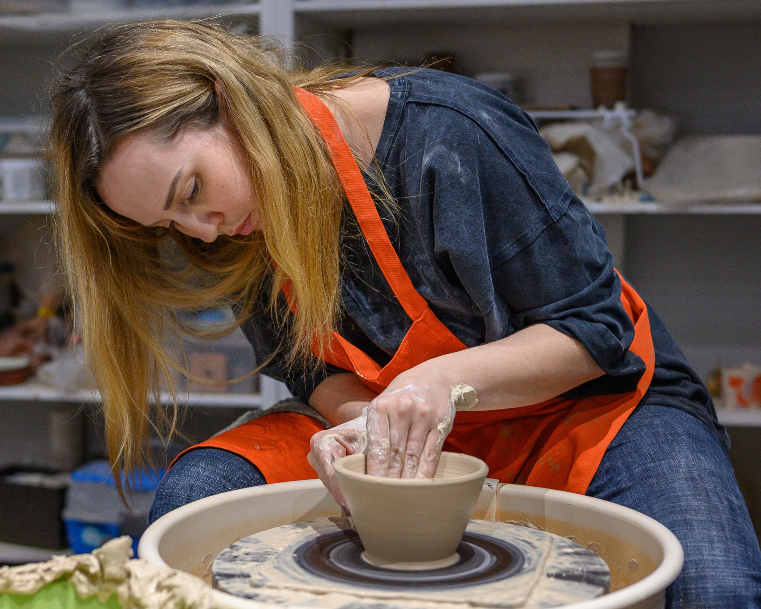 Person in an orange apron shaping a clay bowl on a pottery wheel.