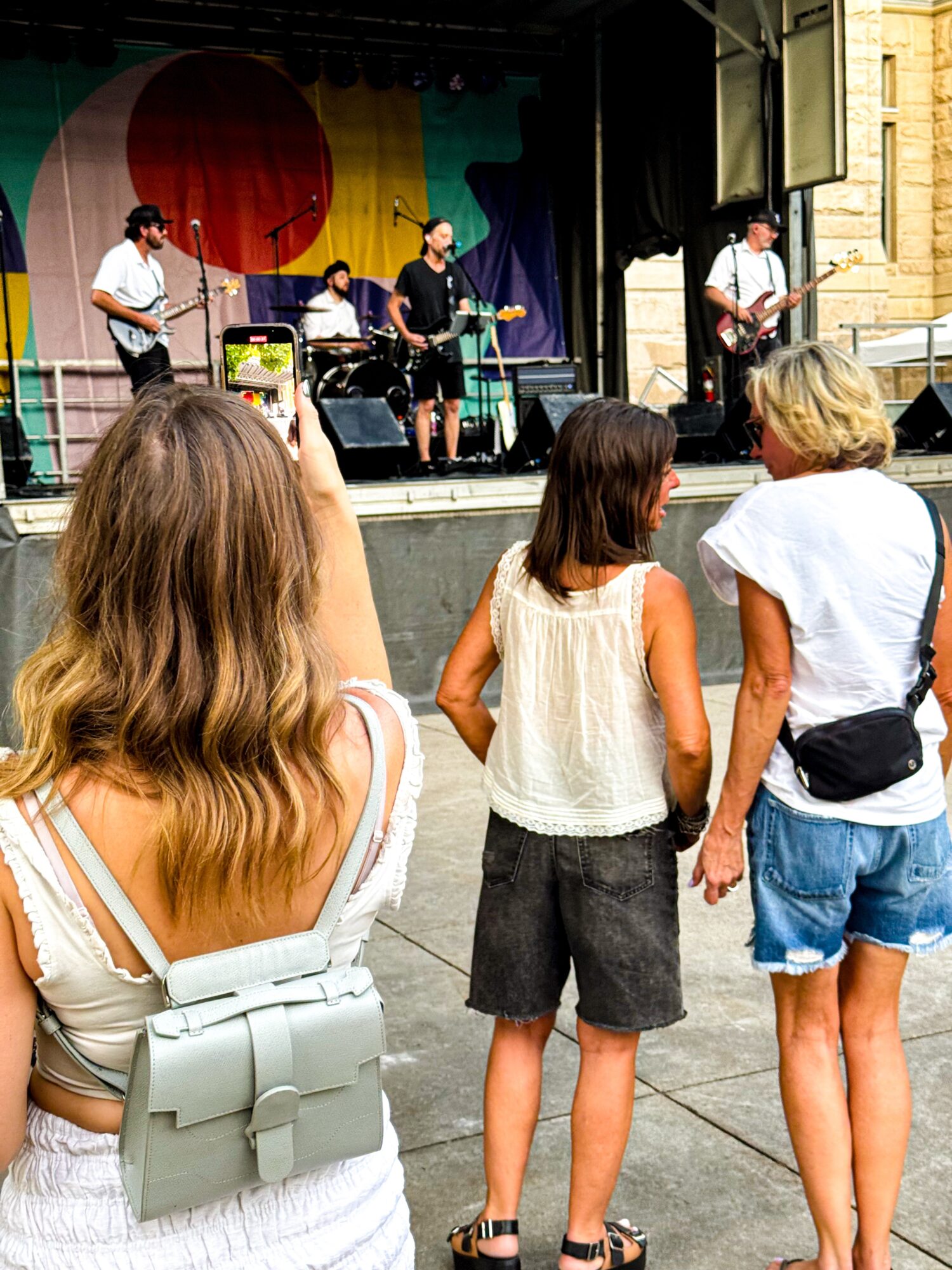 Three people watch a band performing on an outdoor stage.
