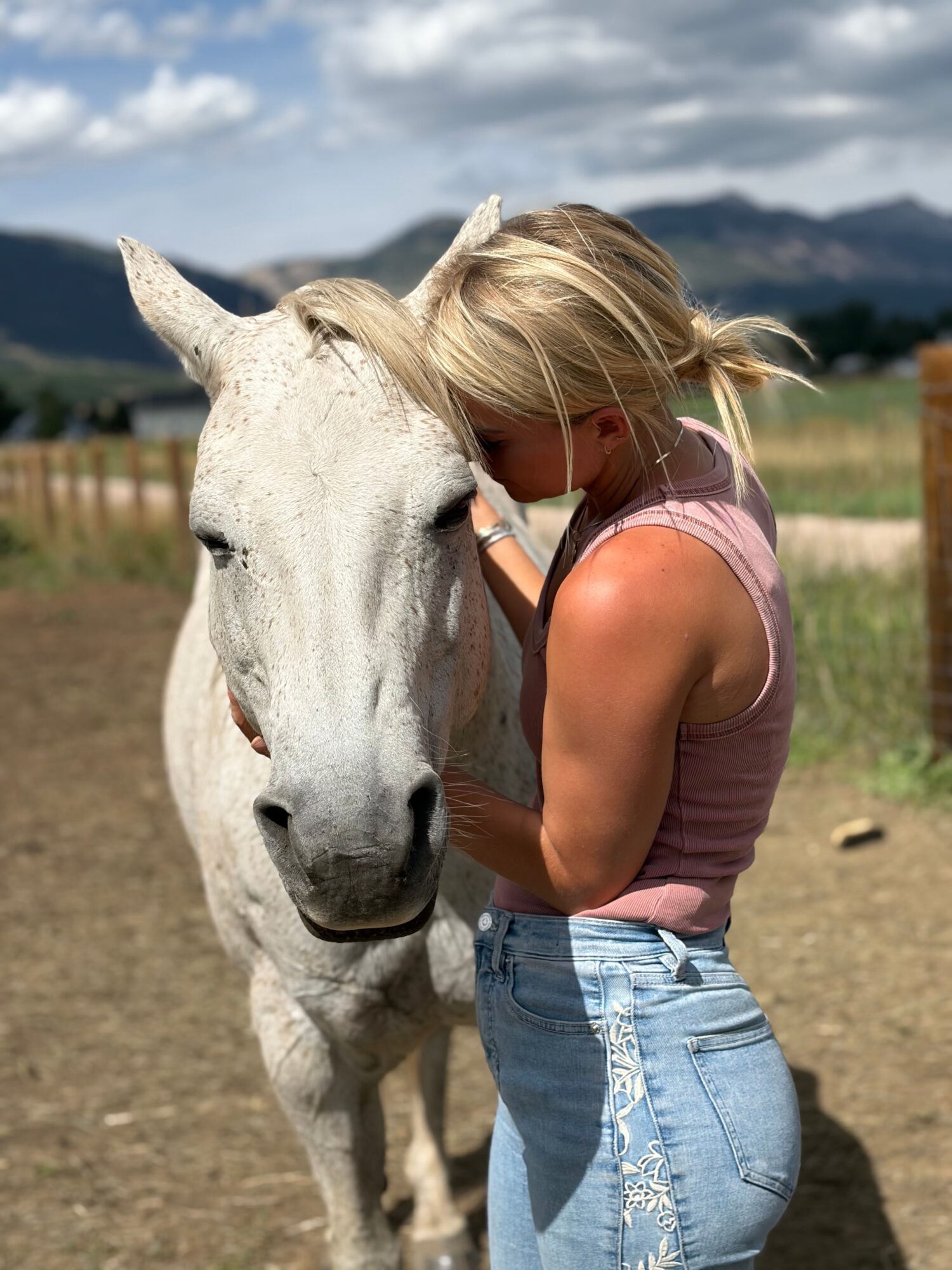 A person leans close to a white horse in a fenced field with mountains in the distance.