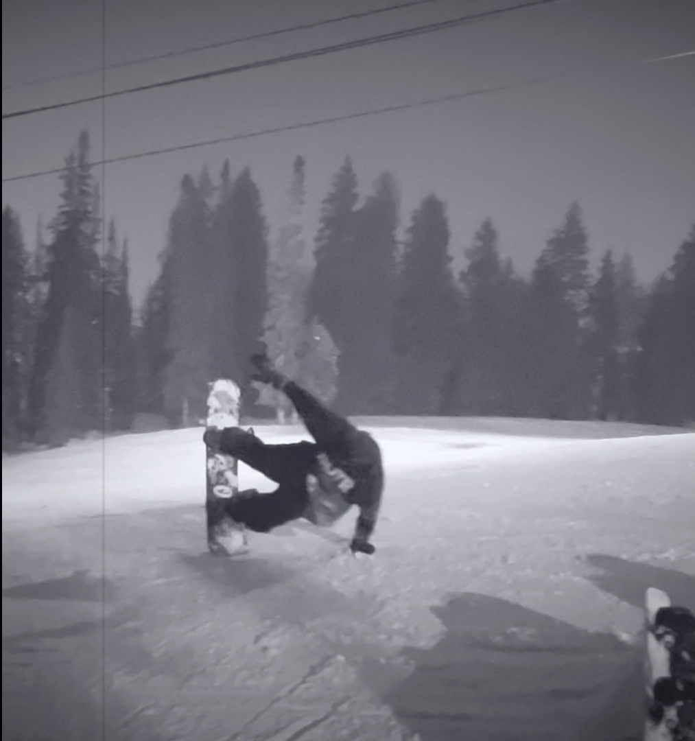 A snowboarder crouches on a snowy slope with trees and overhead cables in the background.