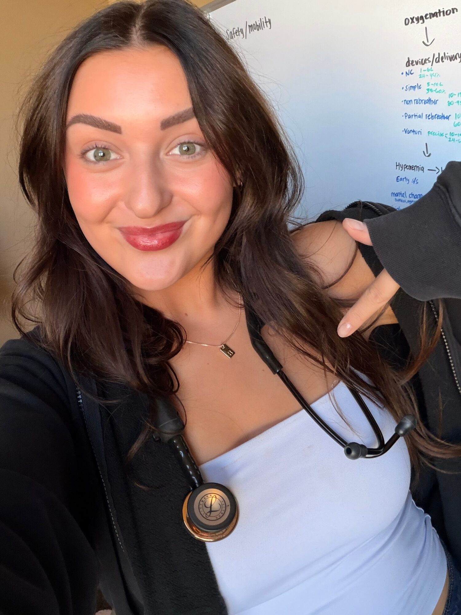 A person in a black cardigan and light blue top poses with a stethoscope in front of a whiteboard.