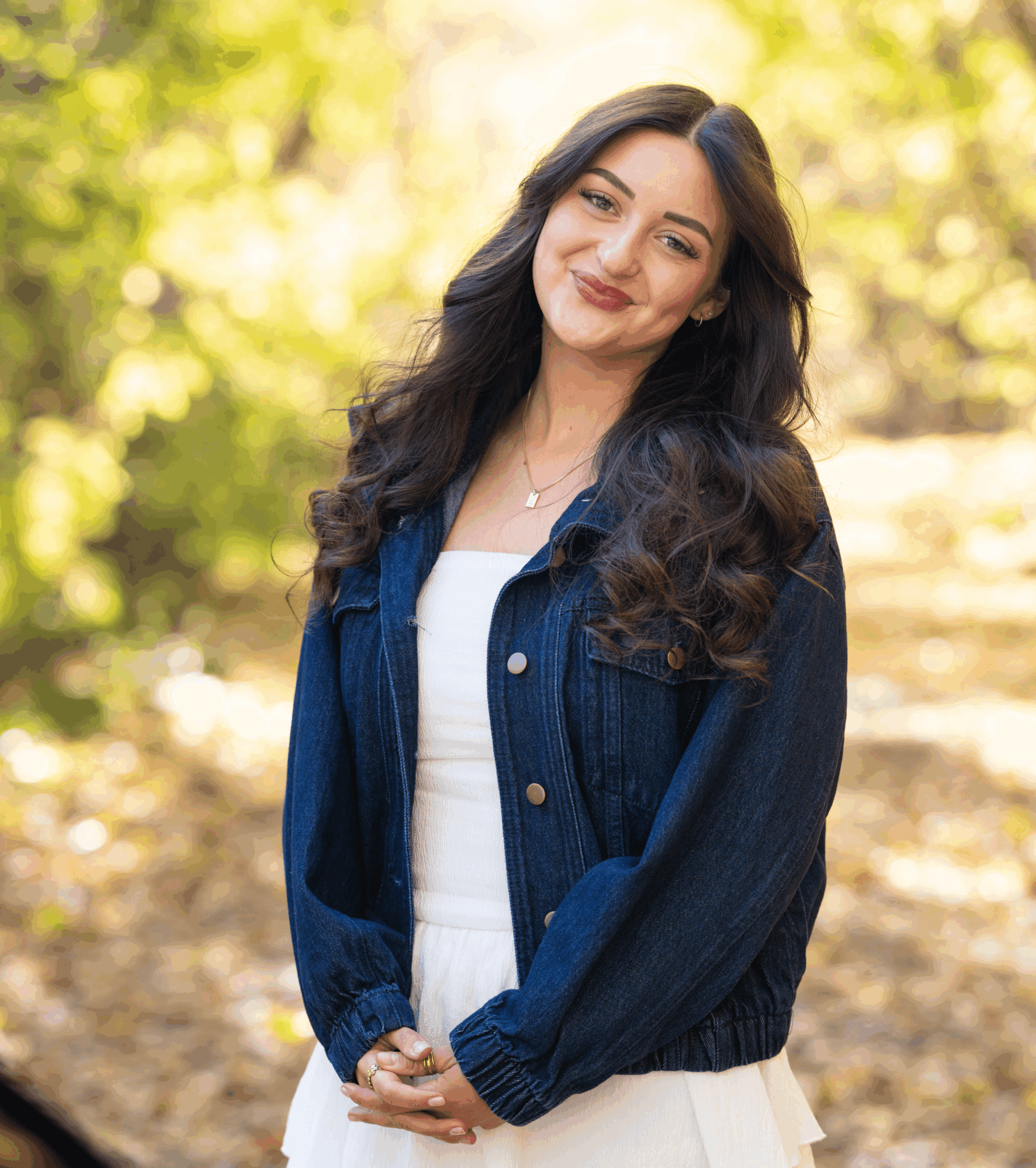 A person with long dark hair stands outdoors in a blue jacket, hands clasped against a sunlit background.