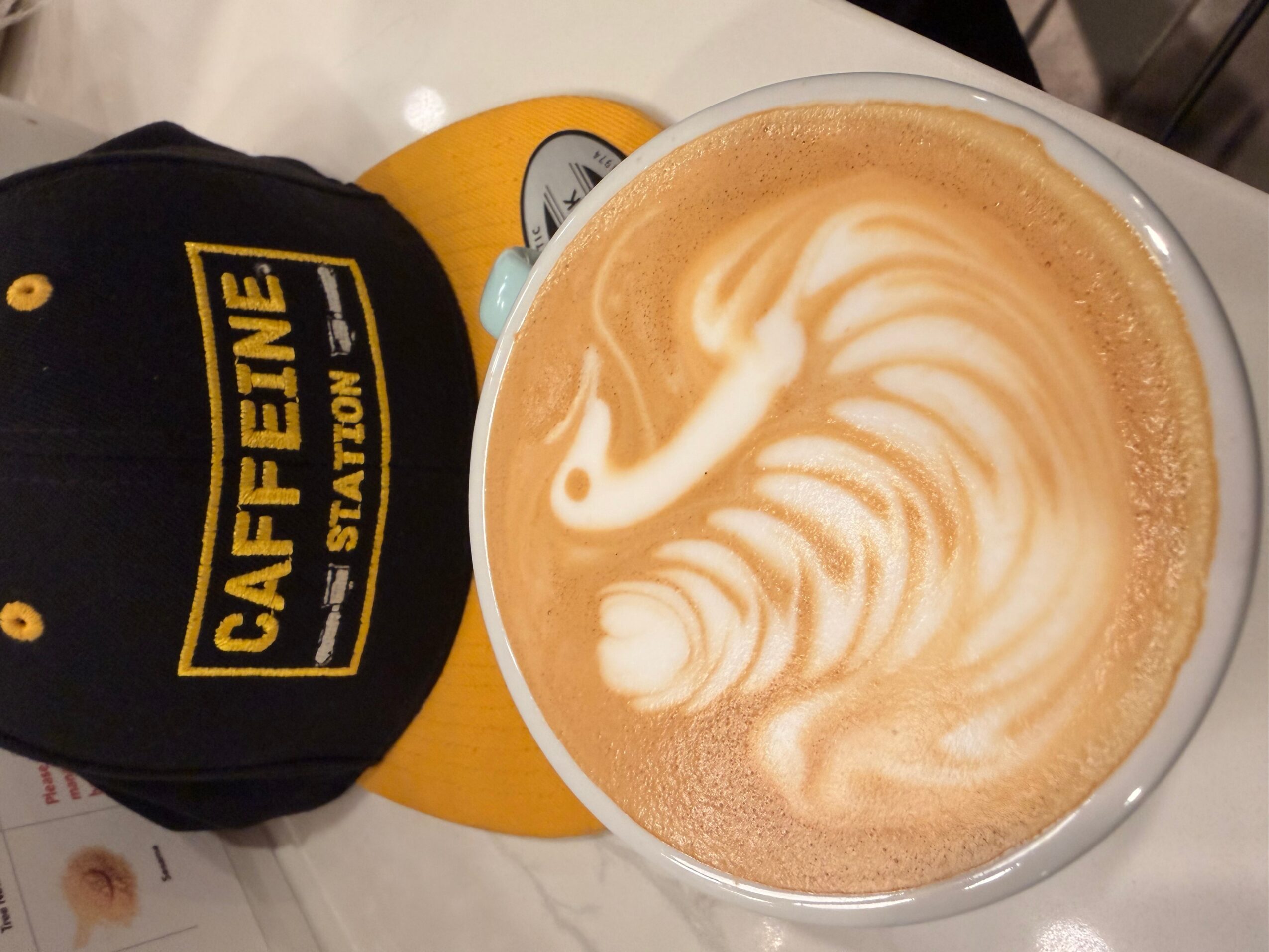 A cup of latte art beside a black cap with yellow “Caffeine Station” lettering.