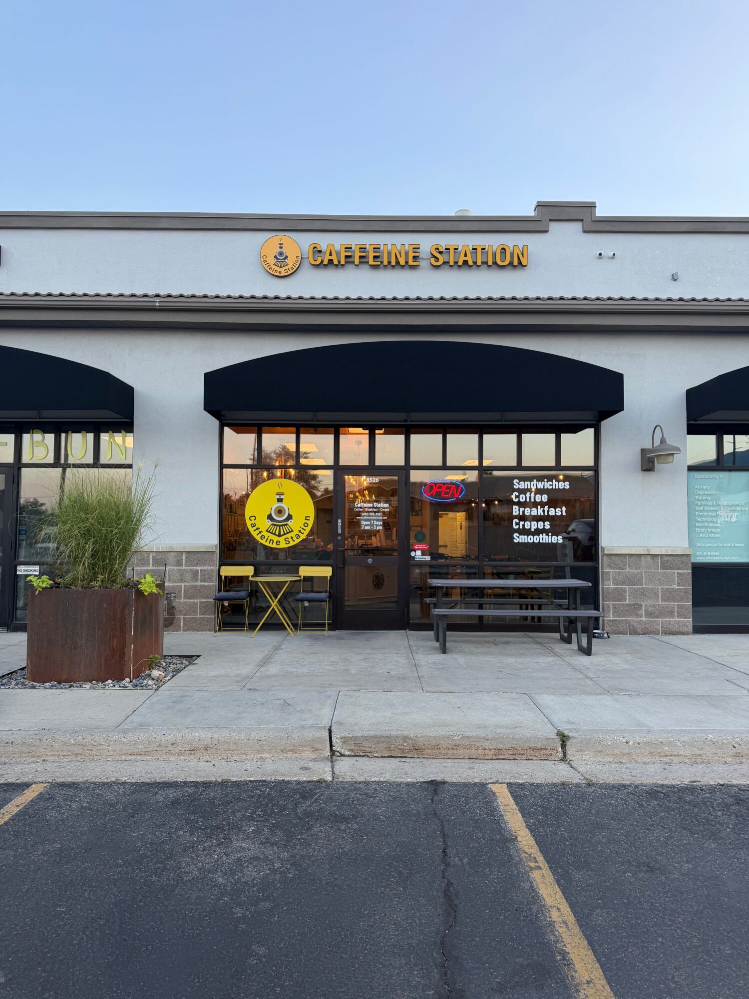 A storefront with a yellow circular sign, dark awnings, glass doors, and a sidewalk beside a parking lot.
