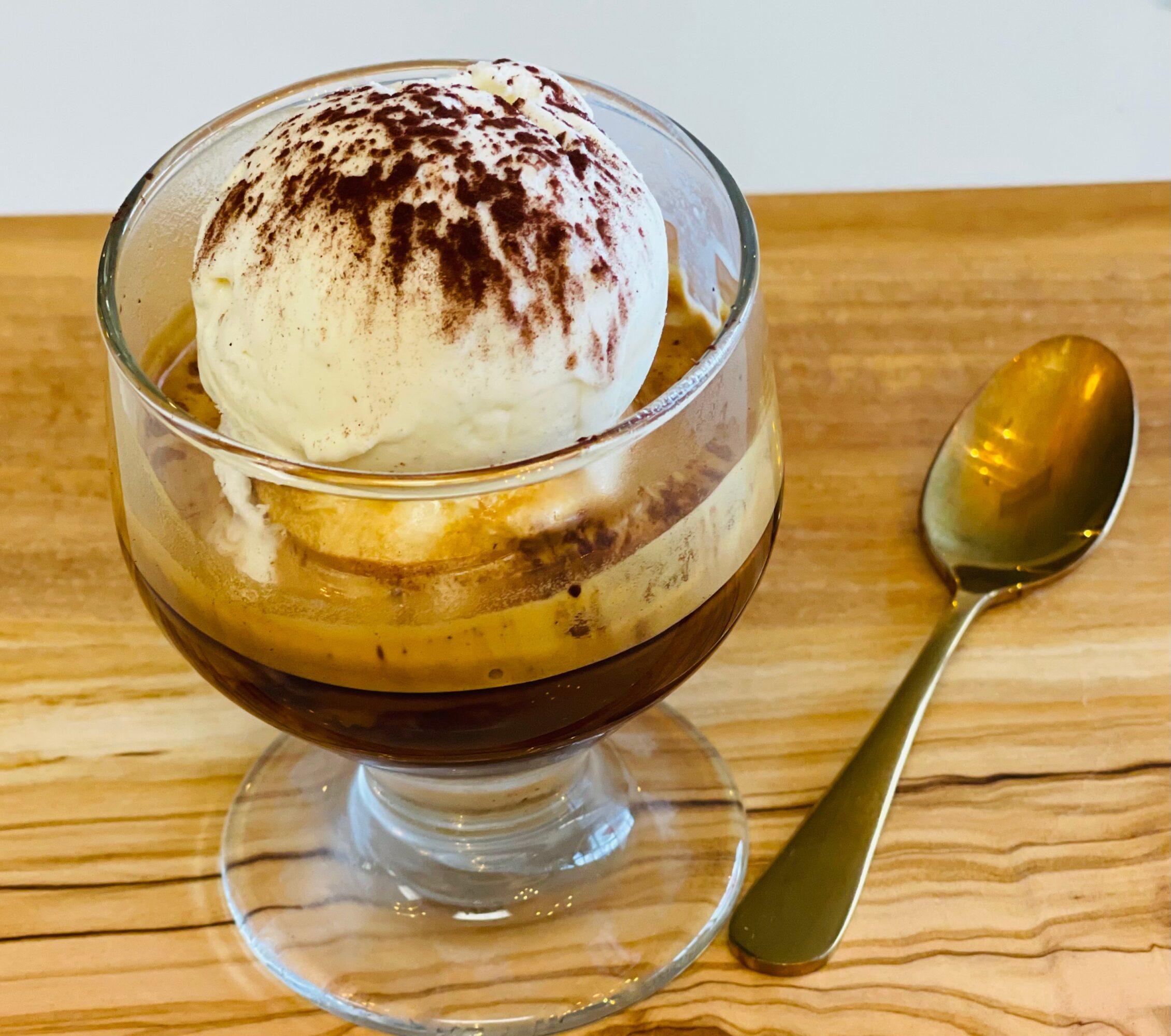 Glass dessert cup with a scoop of cocoa-dusted ice cream, layered with coffee, beside a spoon on a wooden table.