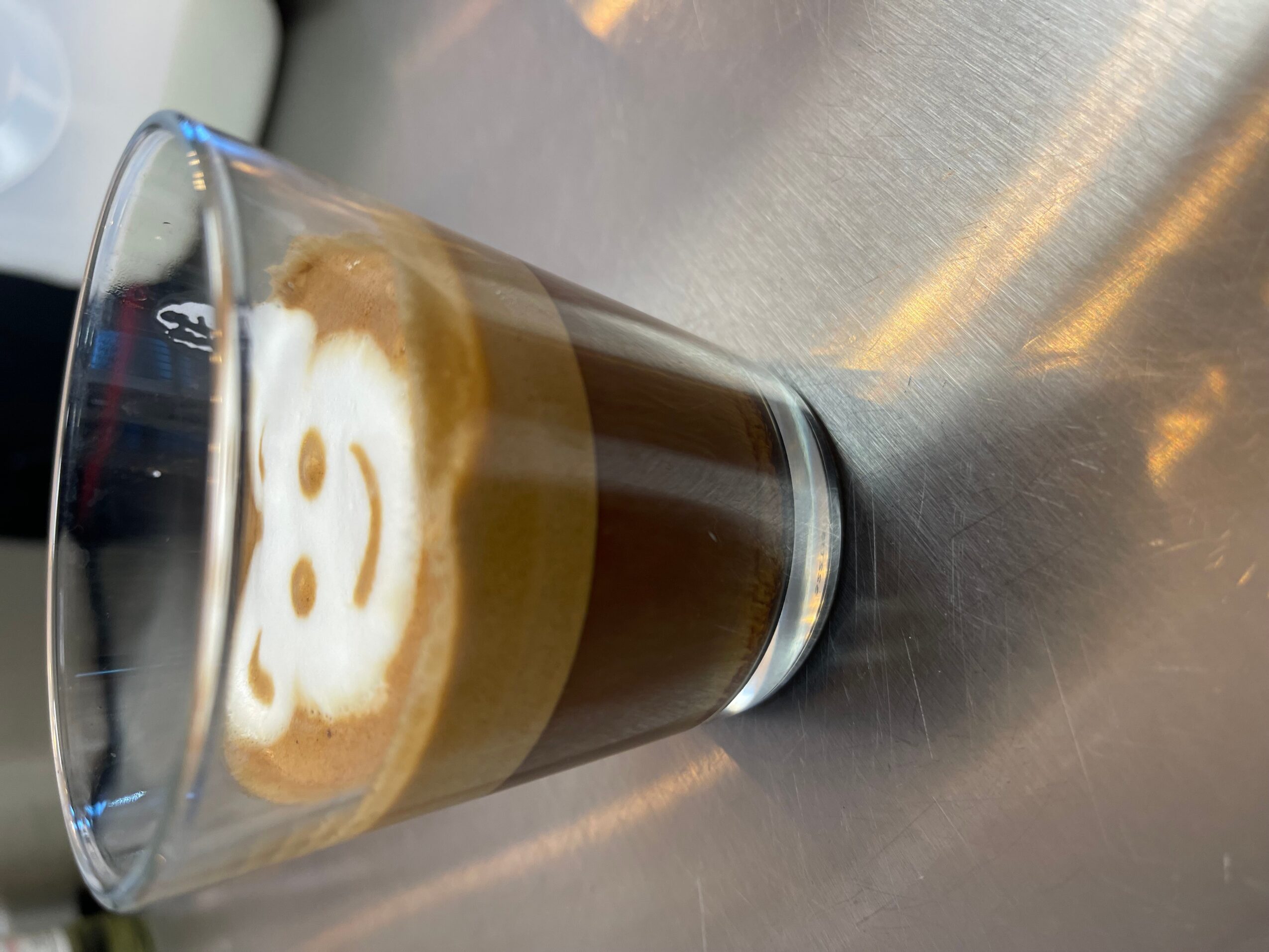 A glass of dark coffee topped with a white foam design on a reflective metal surface.
